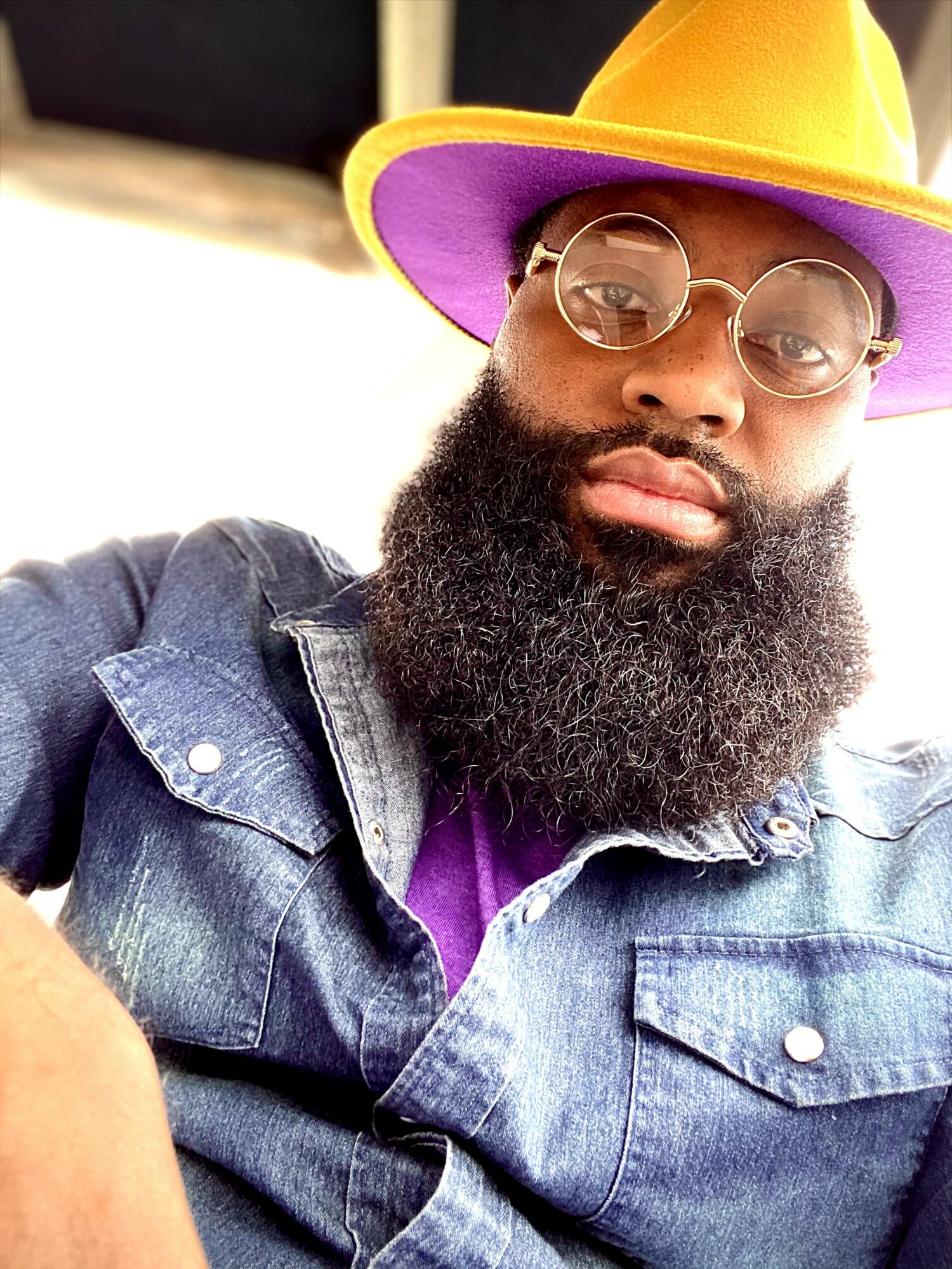 Person with a large beard wearing glasses, a yellow and purple hat, and a denim jacket.