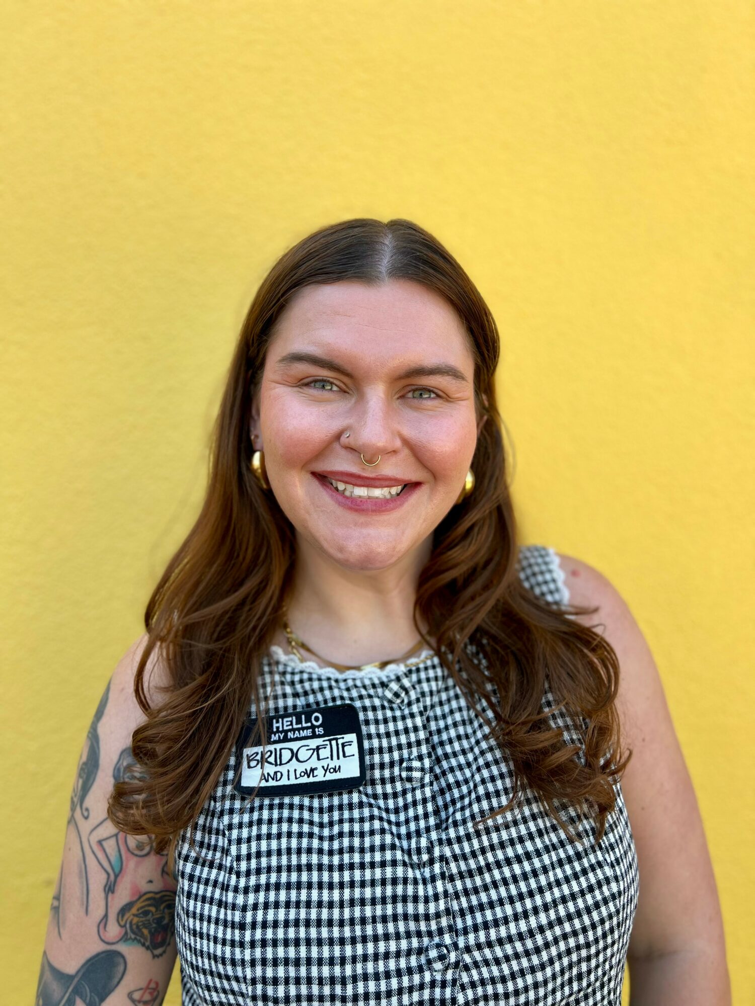 Smiling woman with long brown hair, wearing a checkered top and earrings, standing against a yellow background.