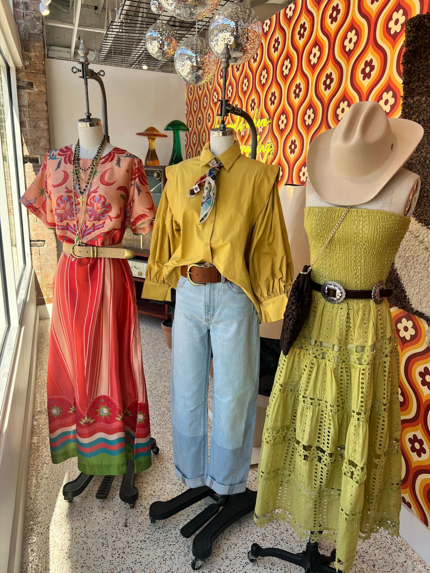 Four mannequins display colorful dresses and outfits in a boutique, with patterned curtains and clothing racks in the background.