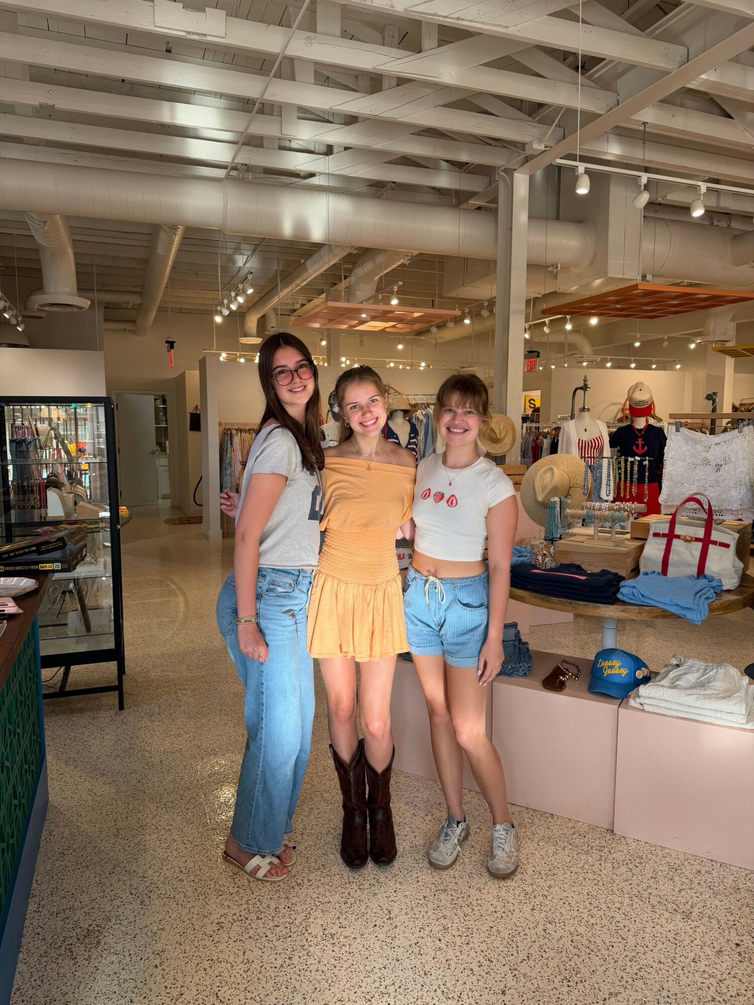 Three women standing together in a shopping mall, with a staircase and store displays in the background.