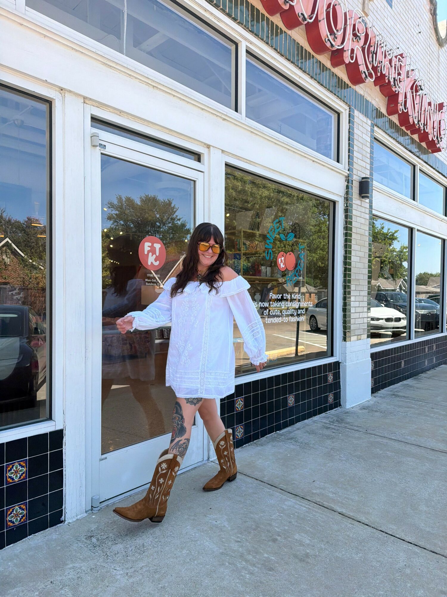 Person in white dress and brown boots standing outside glass door, reflected in window, with trees and building in background.
