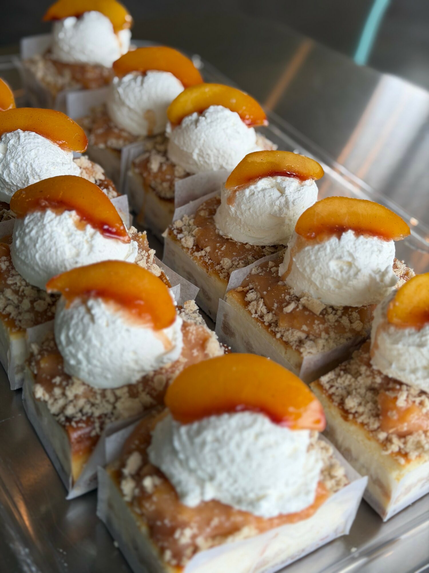 Rows of dessert squares topped with whipped cream and peach slices on a metal tray.