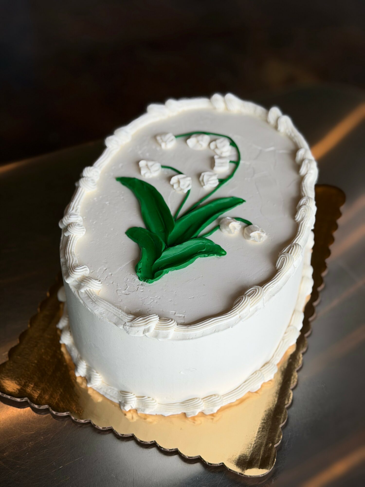 Oval white cake with green leaf decoration and white icing, on a gold-colored base, with a dark background.