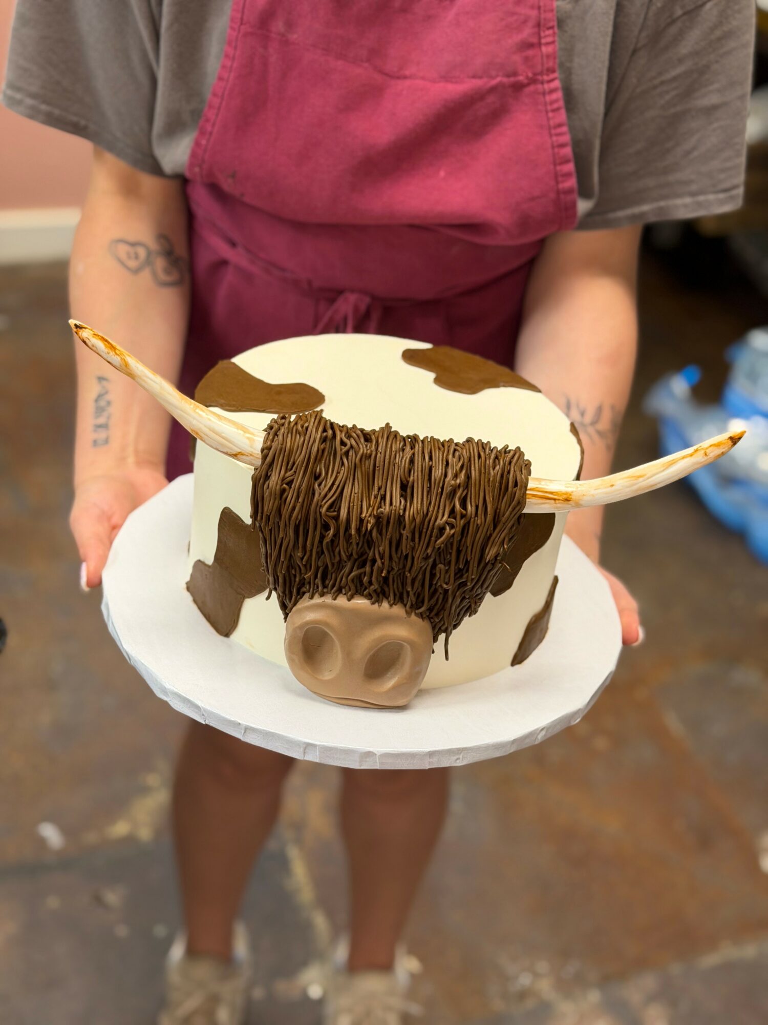 Person holding a cow-shaped cake with horns, ears, and nose, decorated with brown and white icing, on a white plate.
