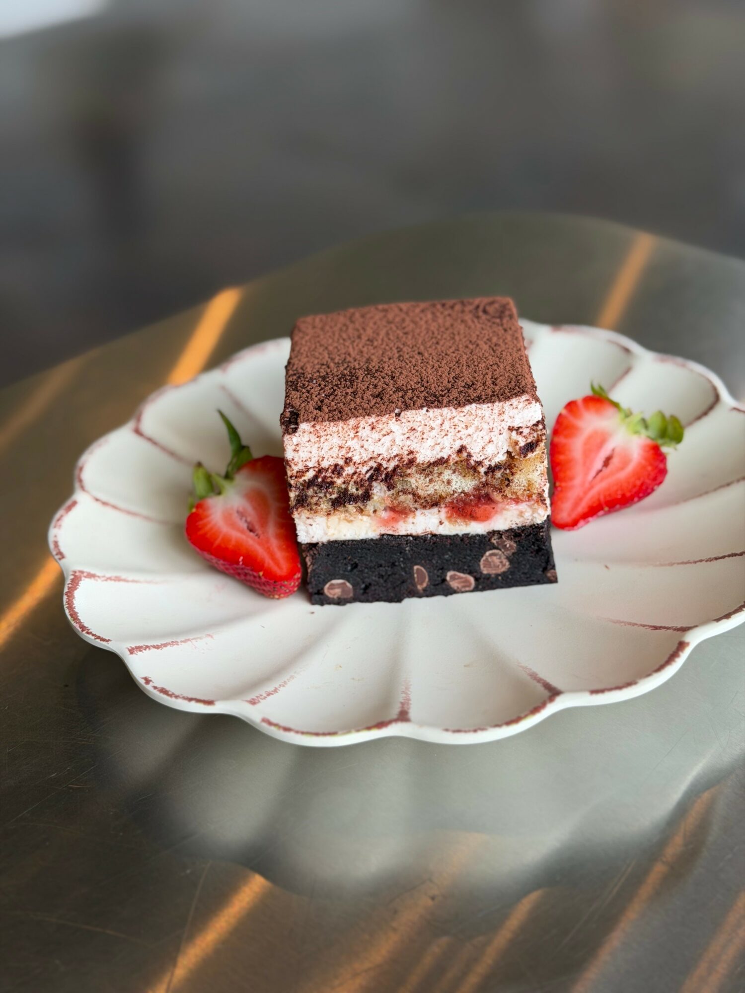 A layered dessert with chocolate, cream, and fruit, garnished with two strawberry halves on a white scalloped plate.