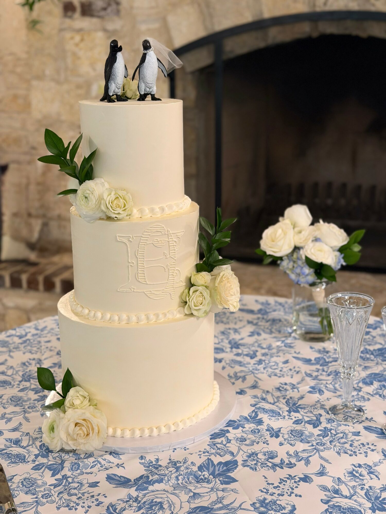 Three-tiered white wedding cake decorated with white roses and greenery, topped with two penguin figurines, on a table with a blue floral tablecloth.