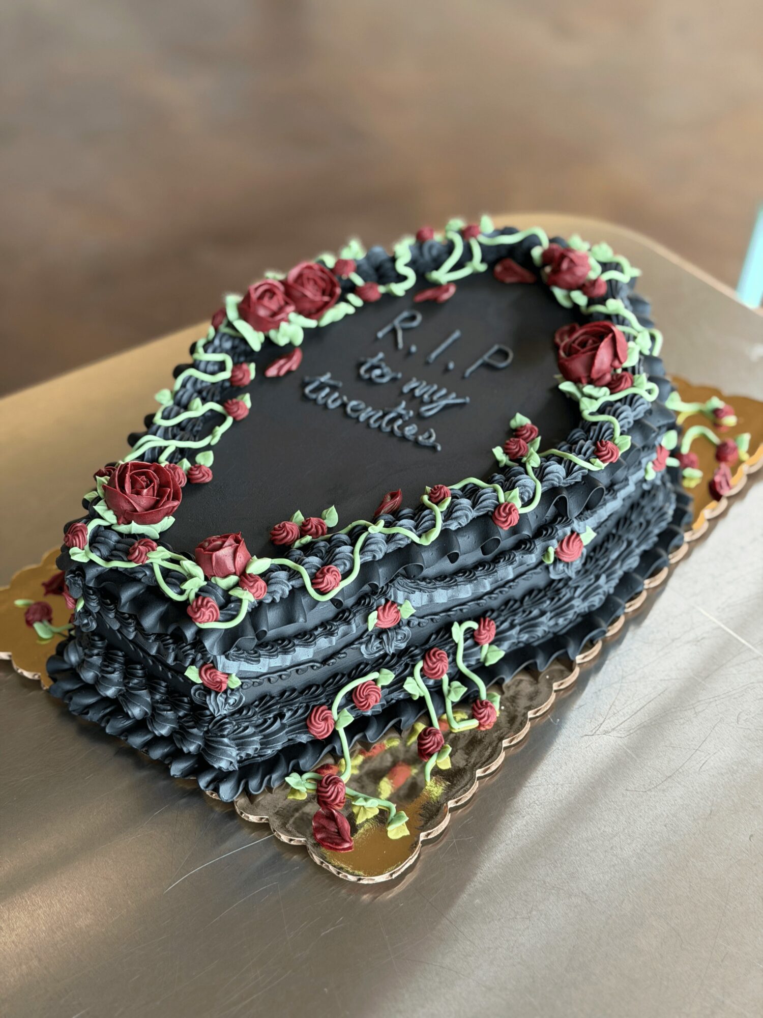 Rectangular black cake with red roses and green icing decorations, with writing on top, on a gold tray.