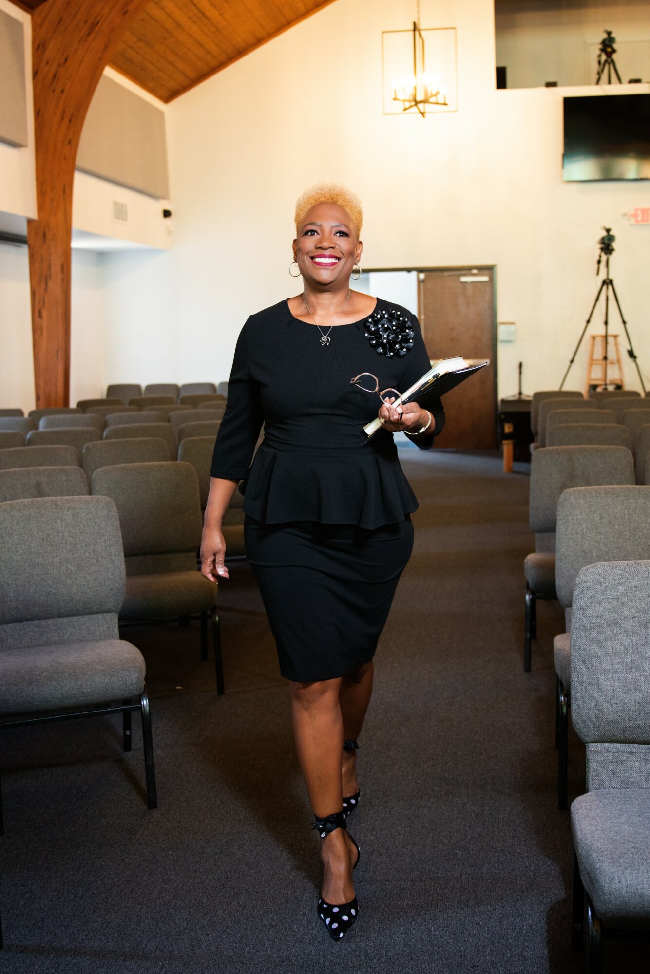 Smiling woman in black dress walking down aisle in church or auditorium, holding a book or folder, with chairs on both sides.