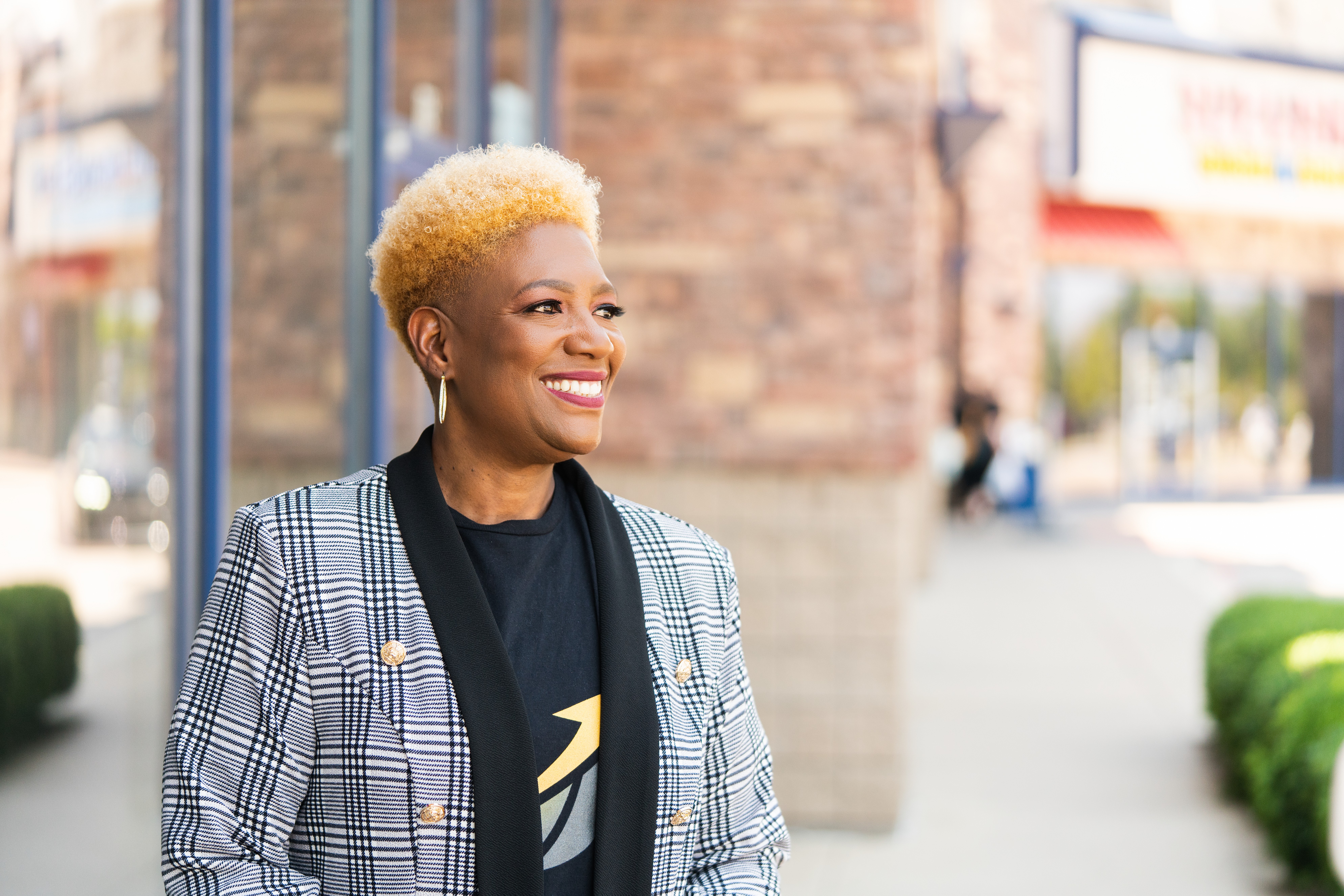 Smiling woman with short blonde hair wearing a plaid blazer and black shirt, standing outdoors in an urban setting.