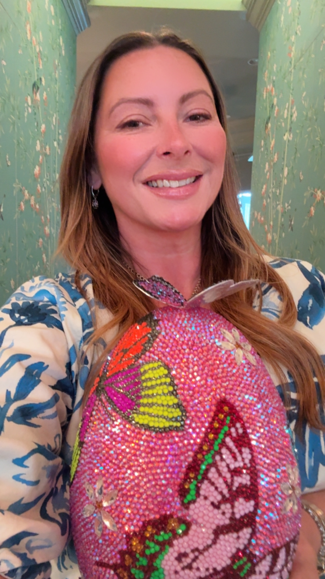 Woman smiling, wearing a colorful, patterned top with a pink background and green and yellow details, in a room with green walls.