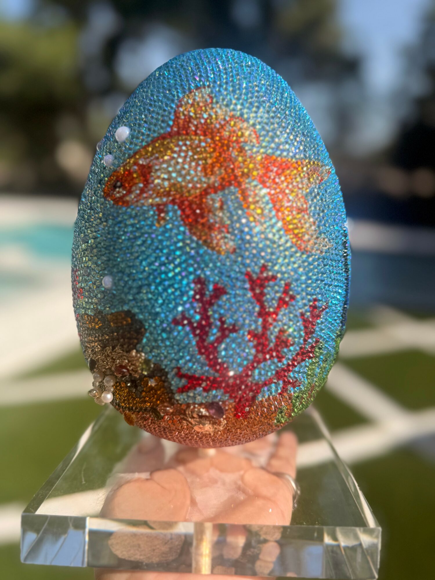 Decorated egg with a goldfish and coral design, covered in small beads, outdoors with blurred background.