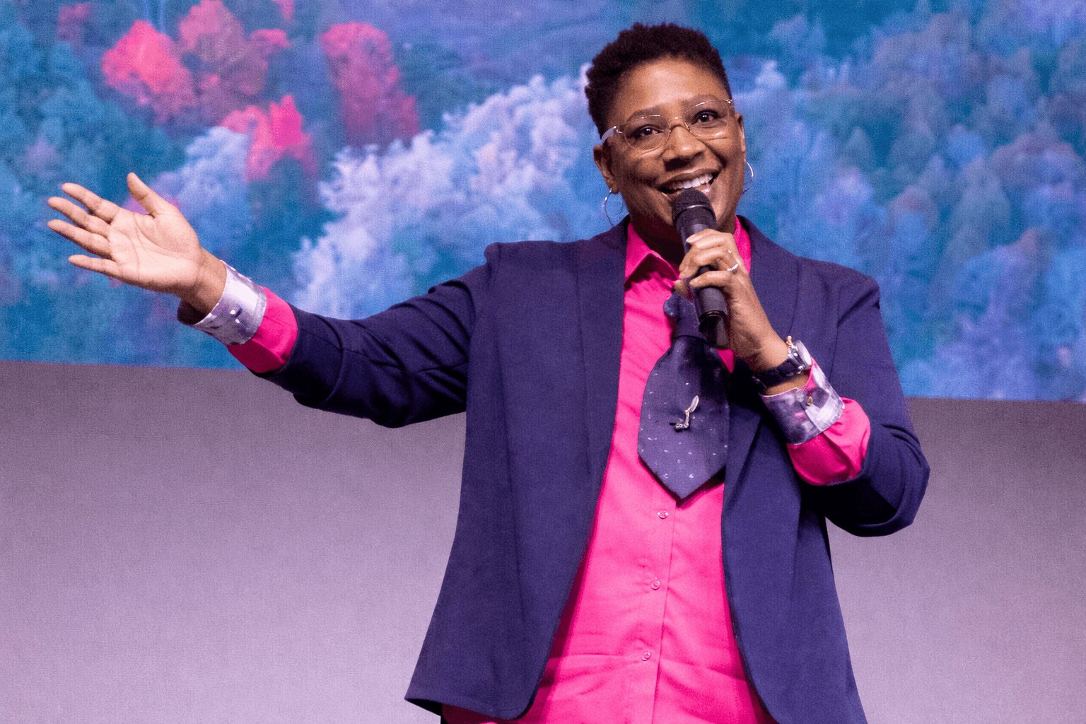 Person speaking into a microphone, smiling, with a colorful cloud background, wearing a pink shirt and dark blazer.