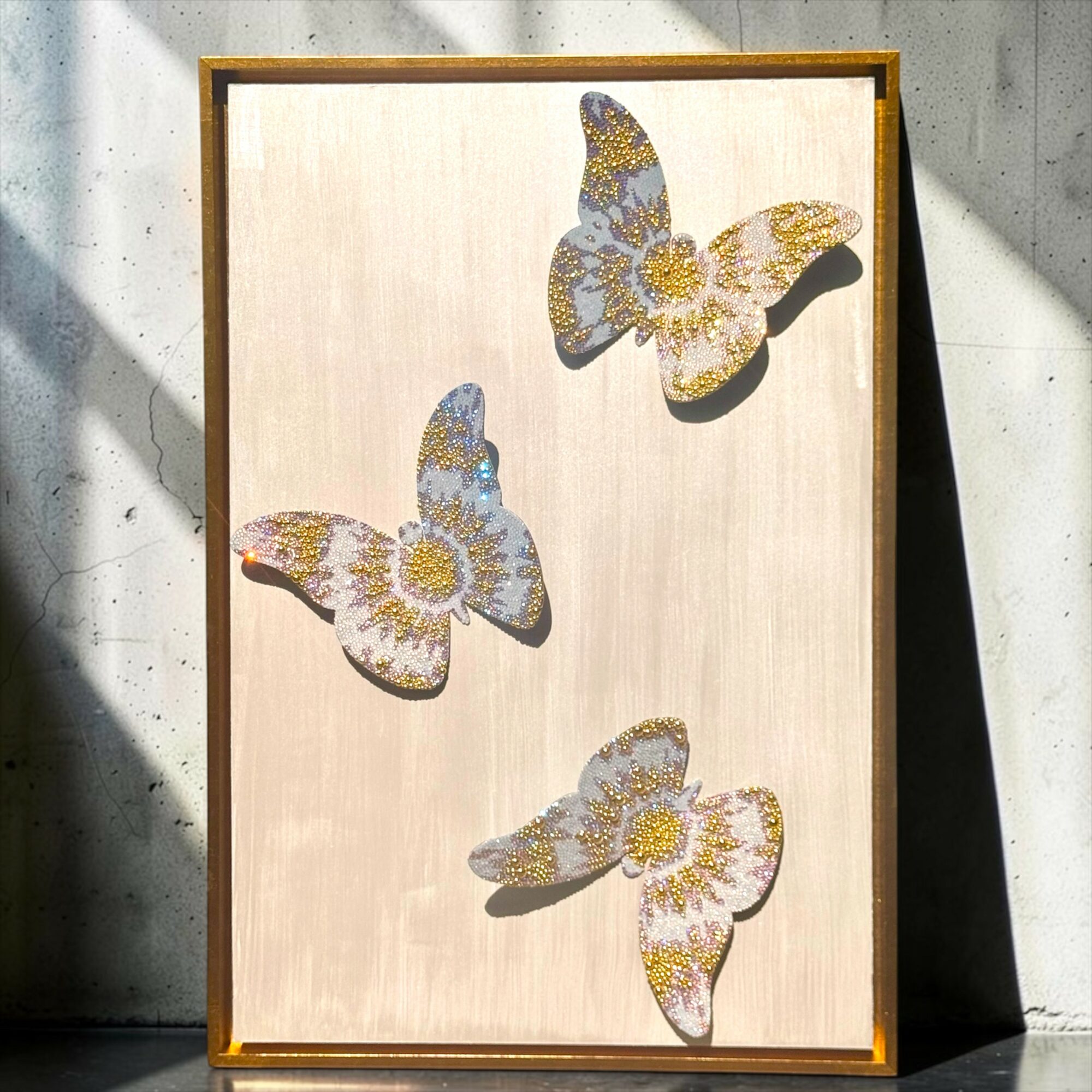 Wooden frame with three butterfly-shaped mosaic art pieces on a light background, leaning against a concrete wall, casting shadows.