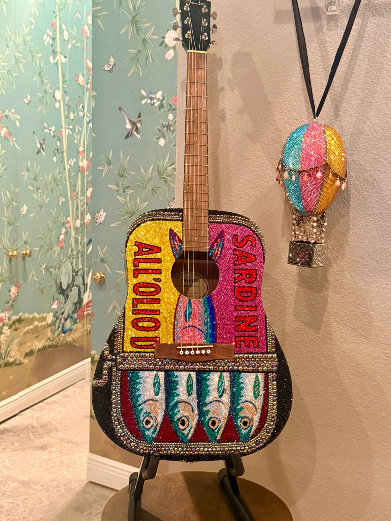 Decorative bag shaped like a guitar with colorful fish and text, hanging on a wall with a beaded ornament nearby.