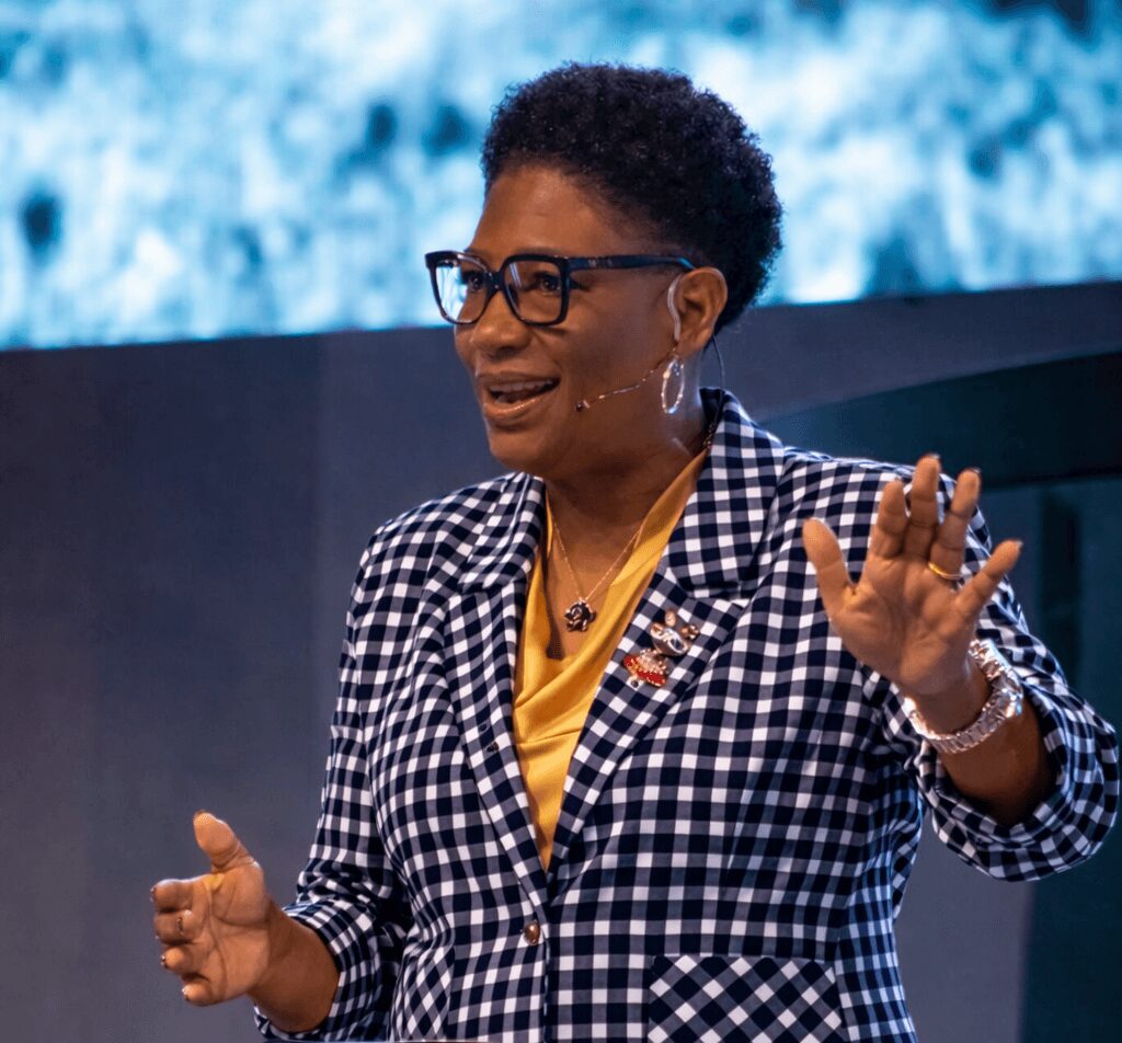 Woman speaking, wearing glasses, a checkered blazer, and a yellow top, gesturing with her hands, in front of a blue background.