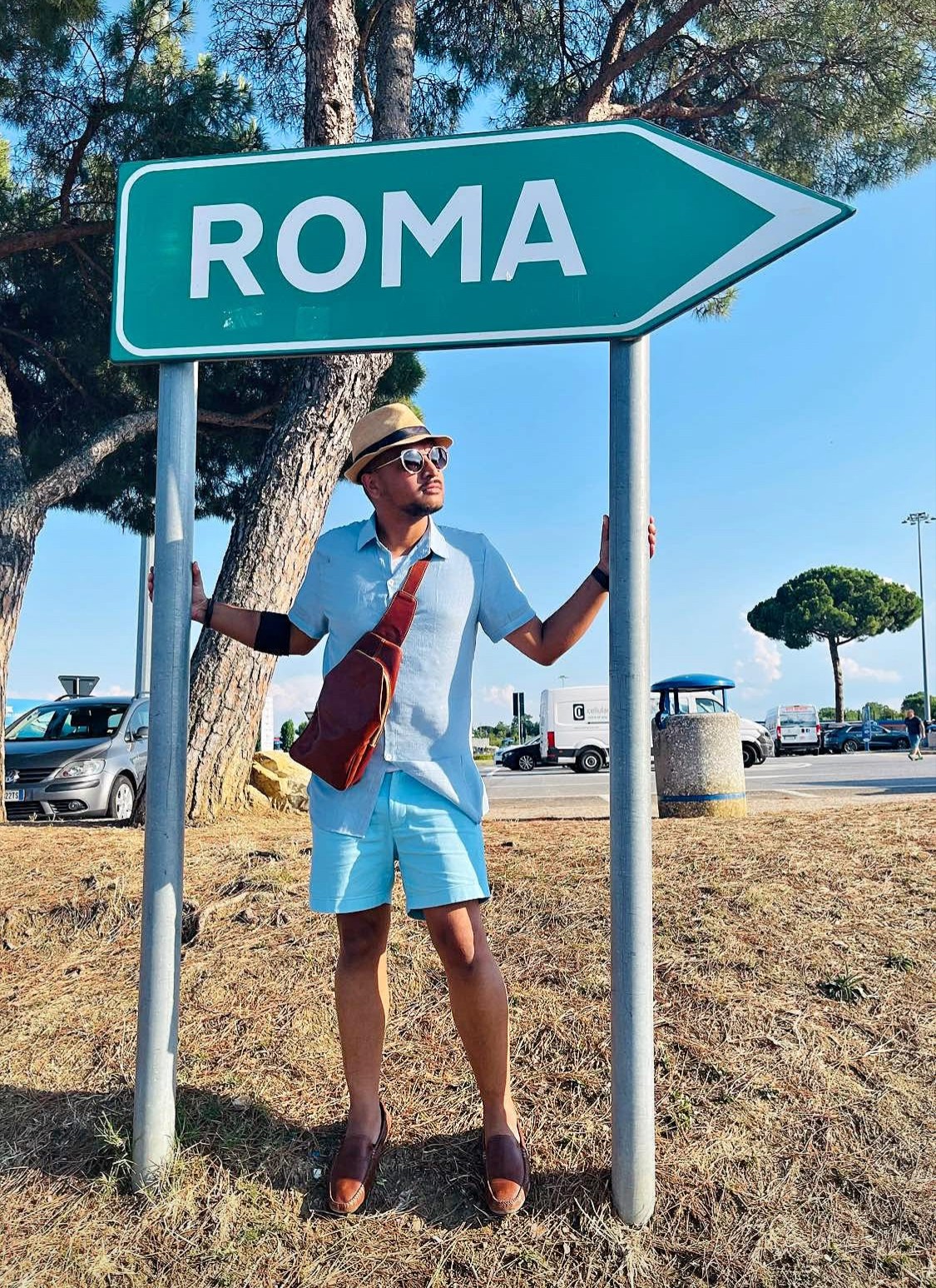 Person standing next to a sign pointing to Rome, wearing sunglasses, a hat, and casual summer clothing, holding the sign post.