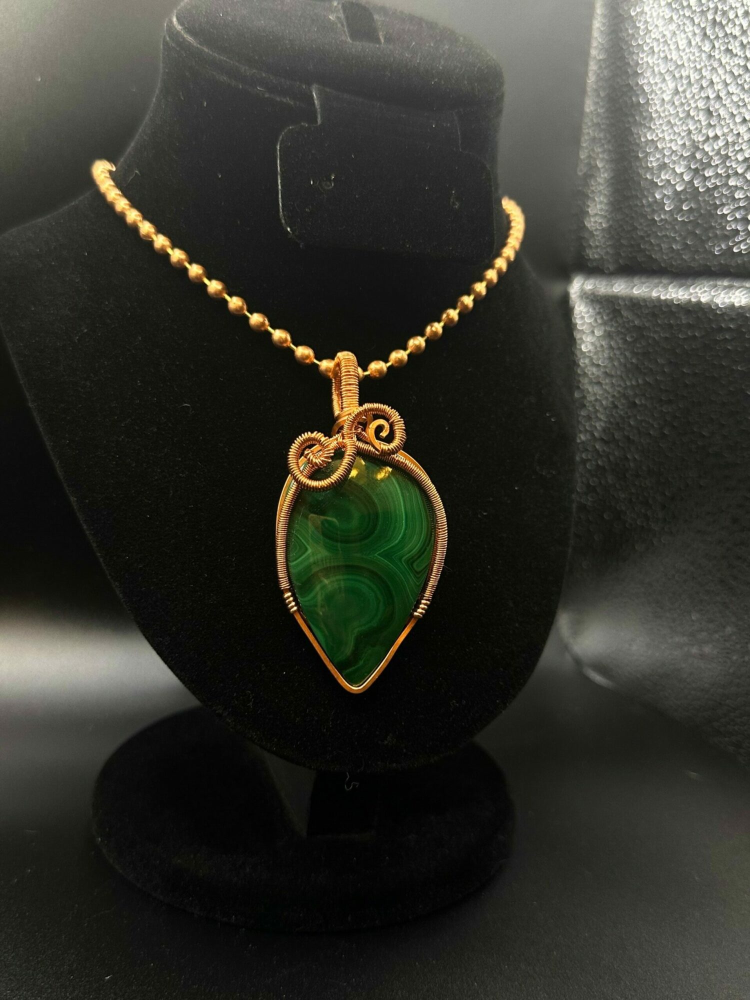 Green gemstone pendant with gold setting on a gold chain, displayed on a black velvet stand.