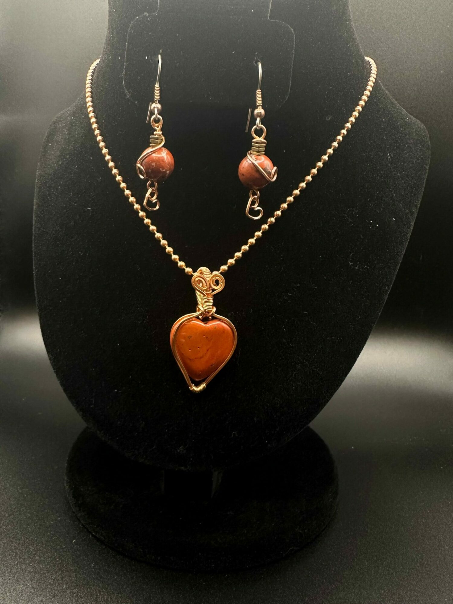 Gold necklace with a heart-shaped pendant and matching earrings displayed on a black stand.