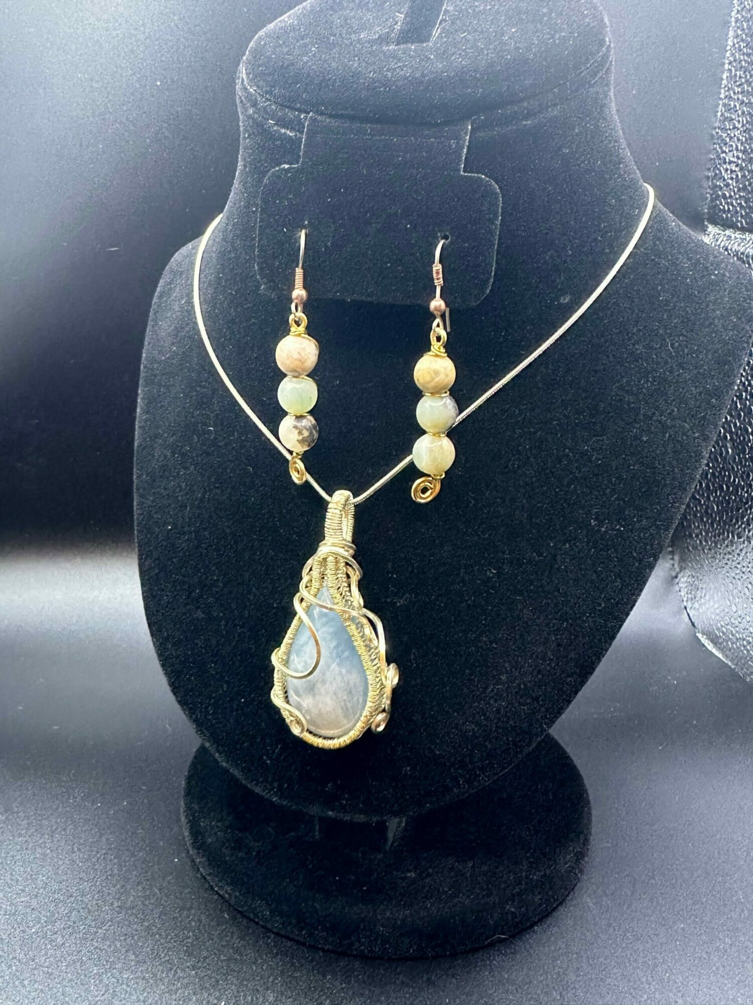 Jewelry displayed on a black velvet bust, including a necklace with beads and a pendant with a stone, against a dark background.