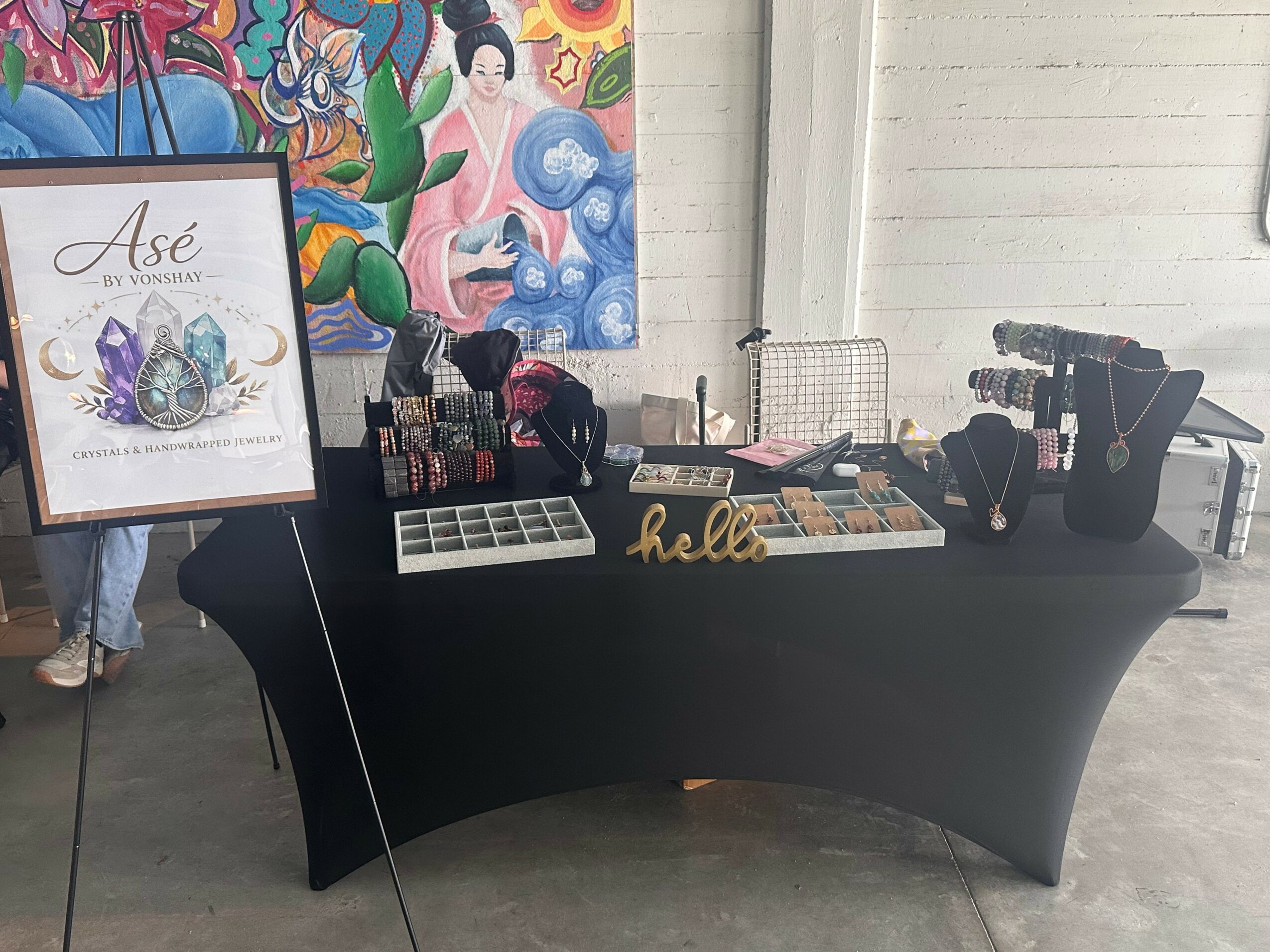 Display table with jewelry, a framed sign, and colorful artwork in background, set against a white wall.