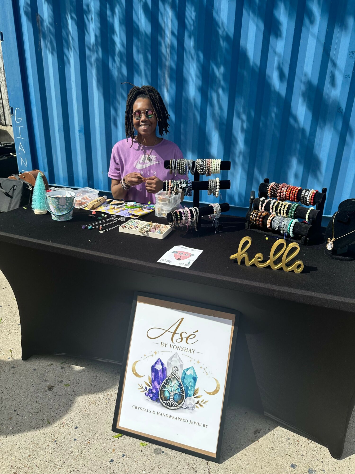 Woman standing behind a table with jewelry and accessories, blue curtain background, black table, framed sign, and sunlight.