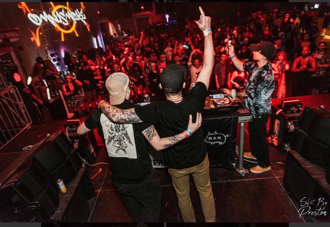 Two people on stage with DJ equipment, facing a large crowd in a nightclub, one raising an arm, colorful lights.