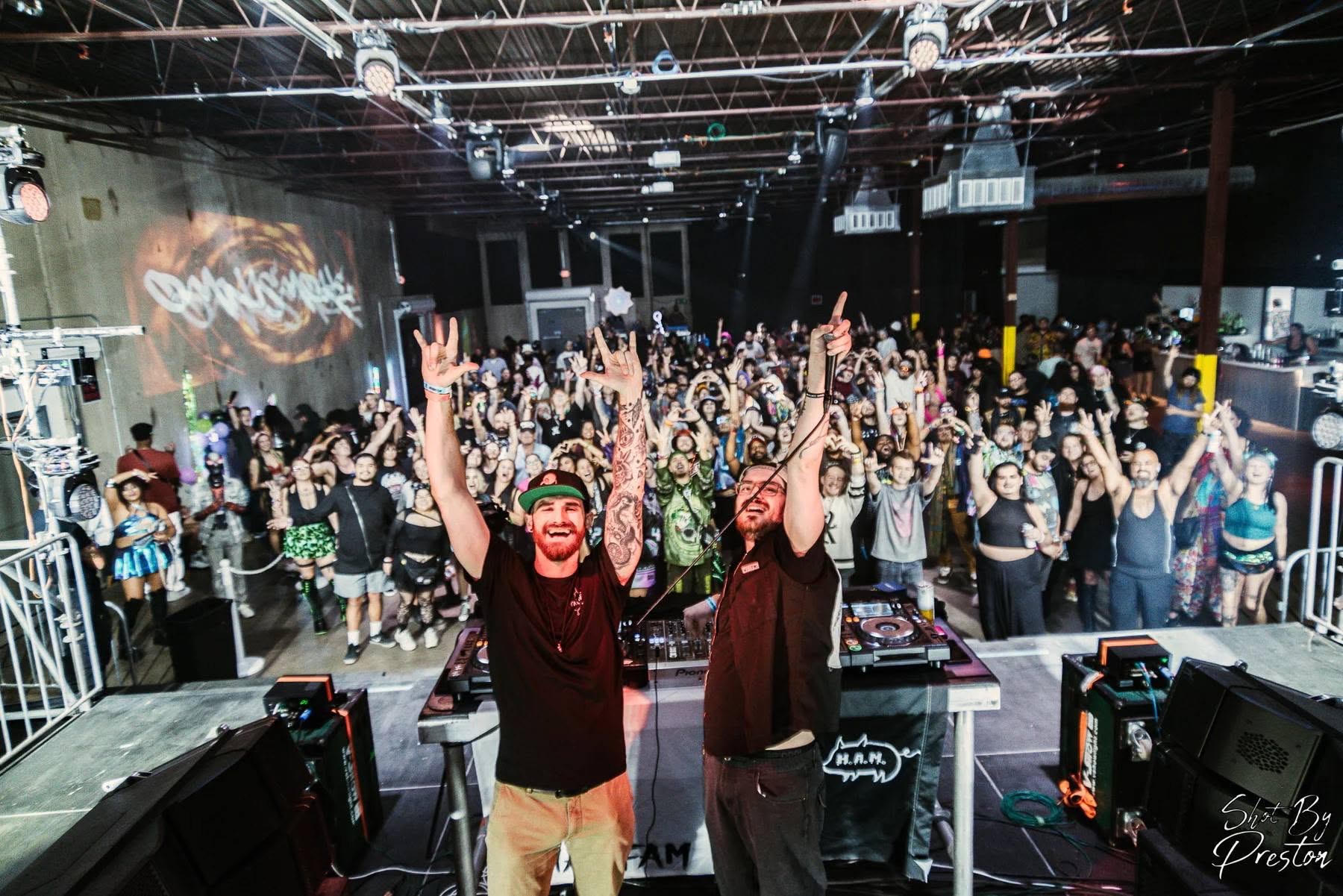 Crowd of people at a concert or event, with some raising their hands, in a large indoor venue with stage lighting.