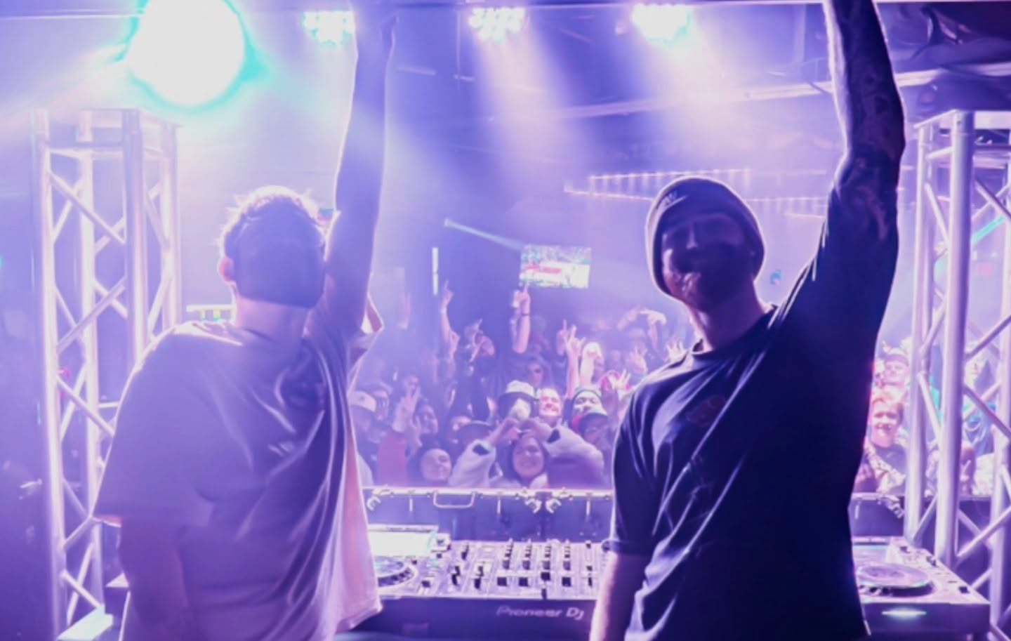 Two DJs perform at a nightclub with a crowd in the background, colorful lighting, and DJ equipment visible.