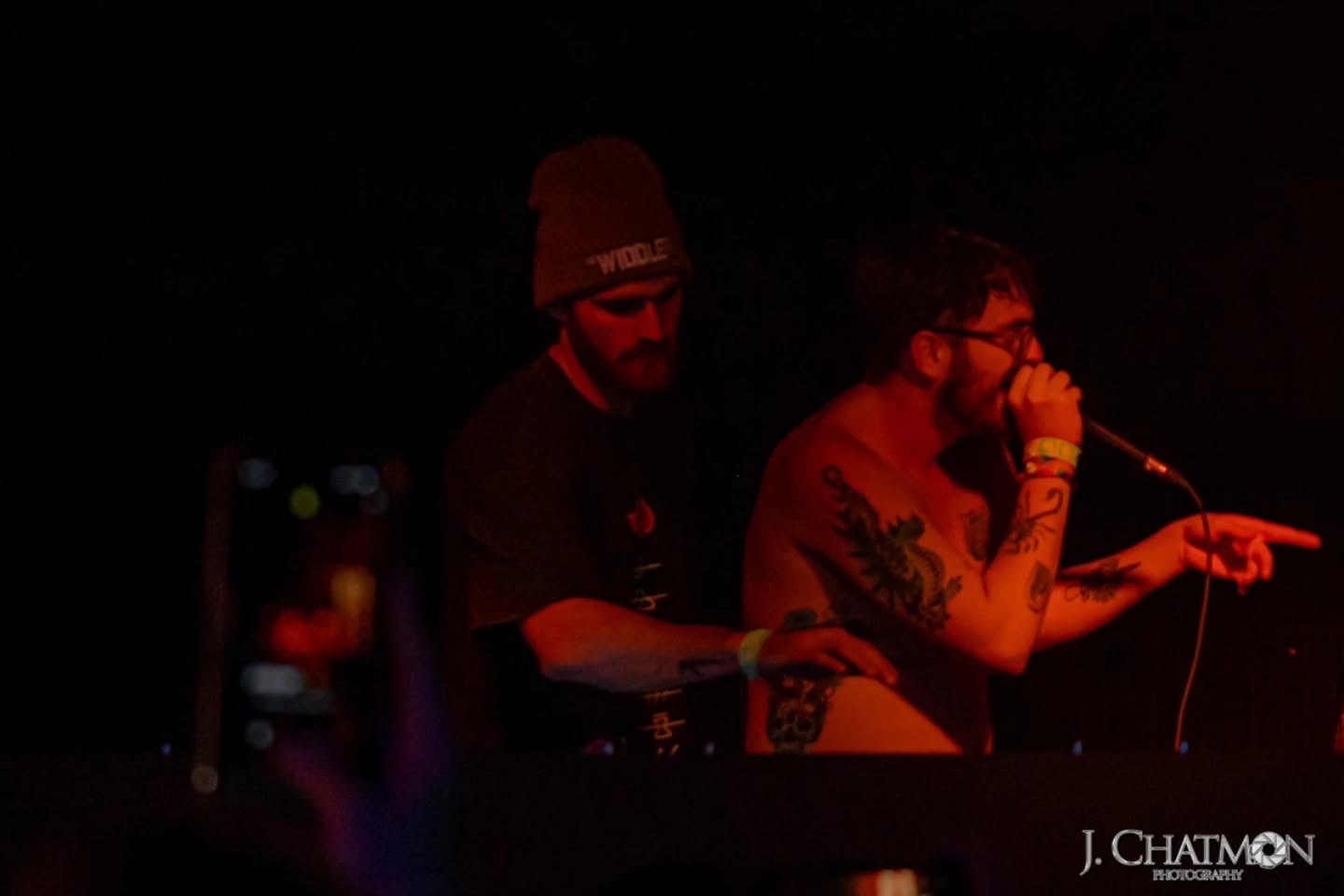 Two people on stage, one with glasses and tattoos, speaking into a microphone, with a dark background.