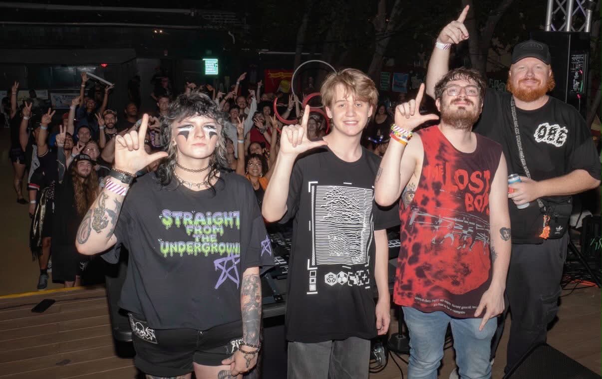 Four people standing together at a concert, making hand gestures, with a crowd in the background.
