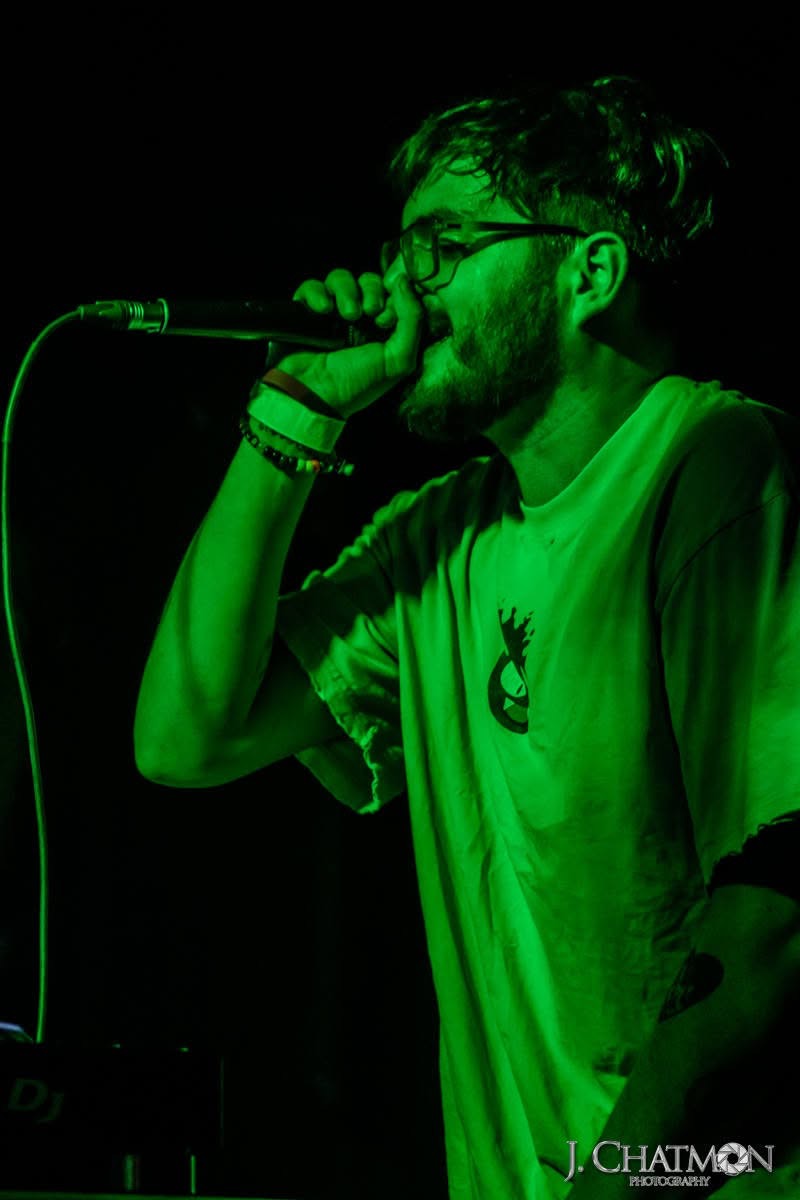 Person singing into microphone on stage, wearing glasses and a loose shirt, illuminated by green light.