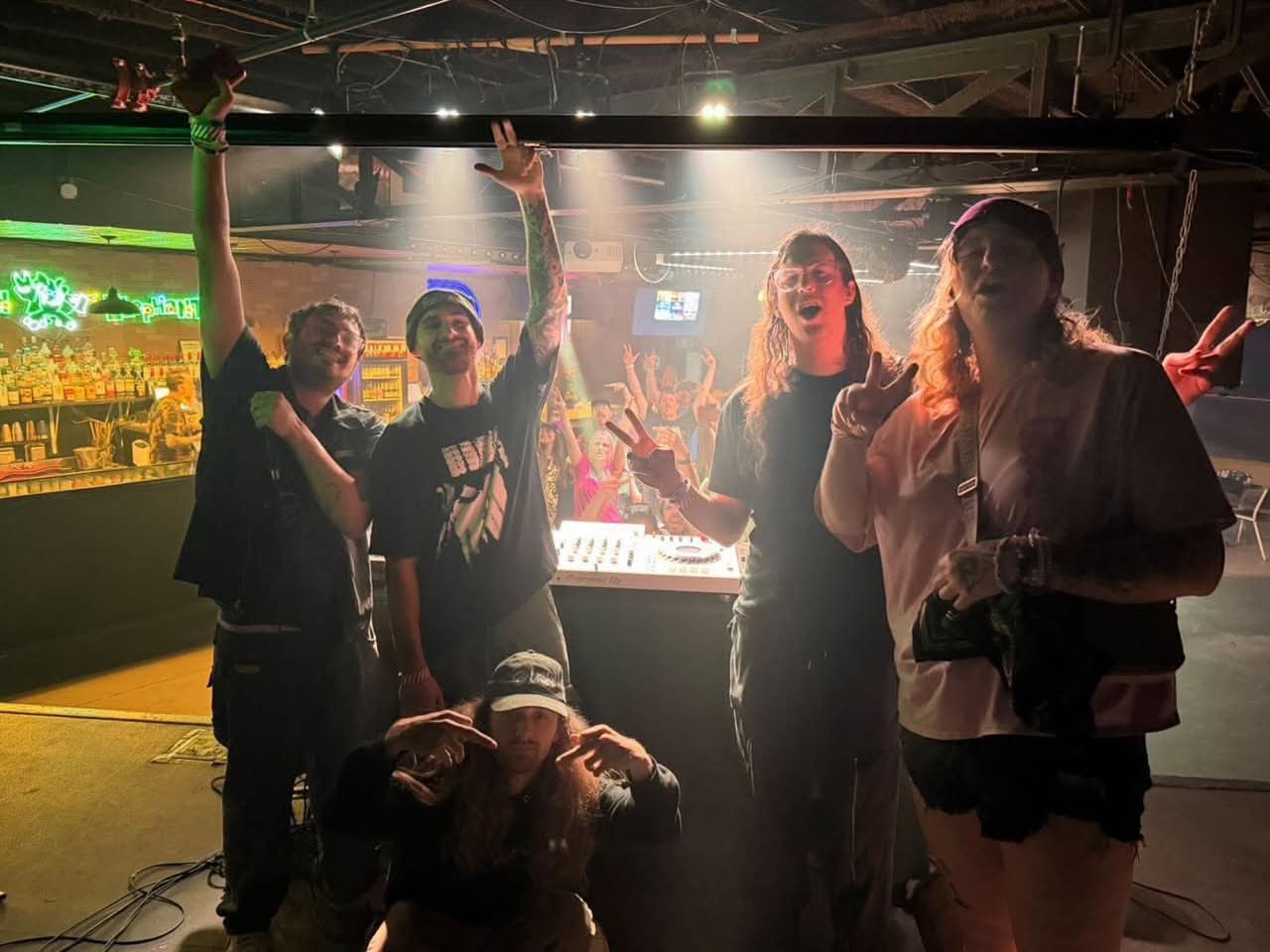 Group of five people posing in a dimly lit bar or club, some making peace signs, with a bar and lights in the background.