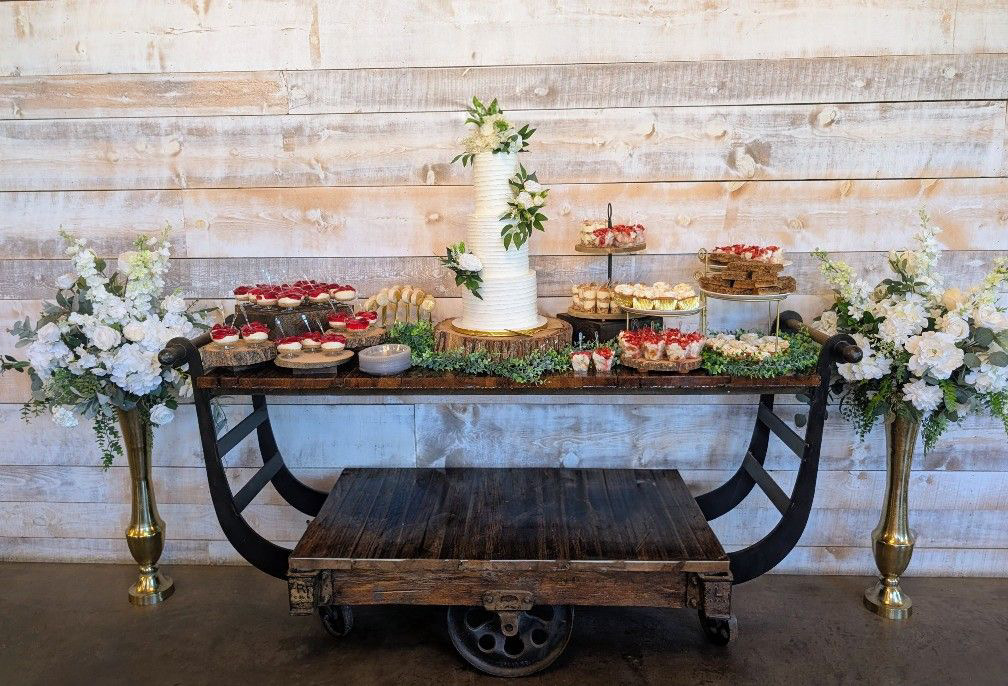 Dessert table with a cake, cupcakes, and fruit, decorated with flowers on a rustic wooden cart against a white wall.