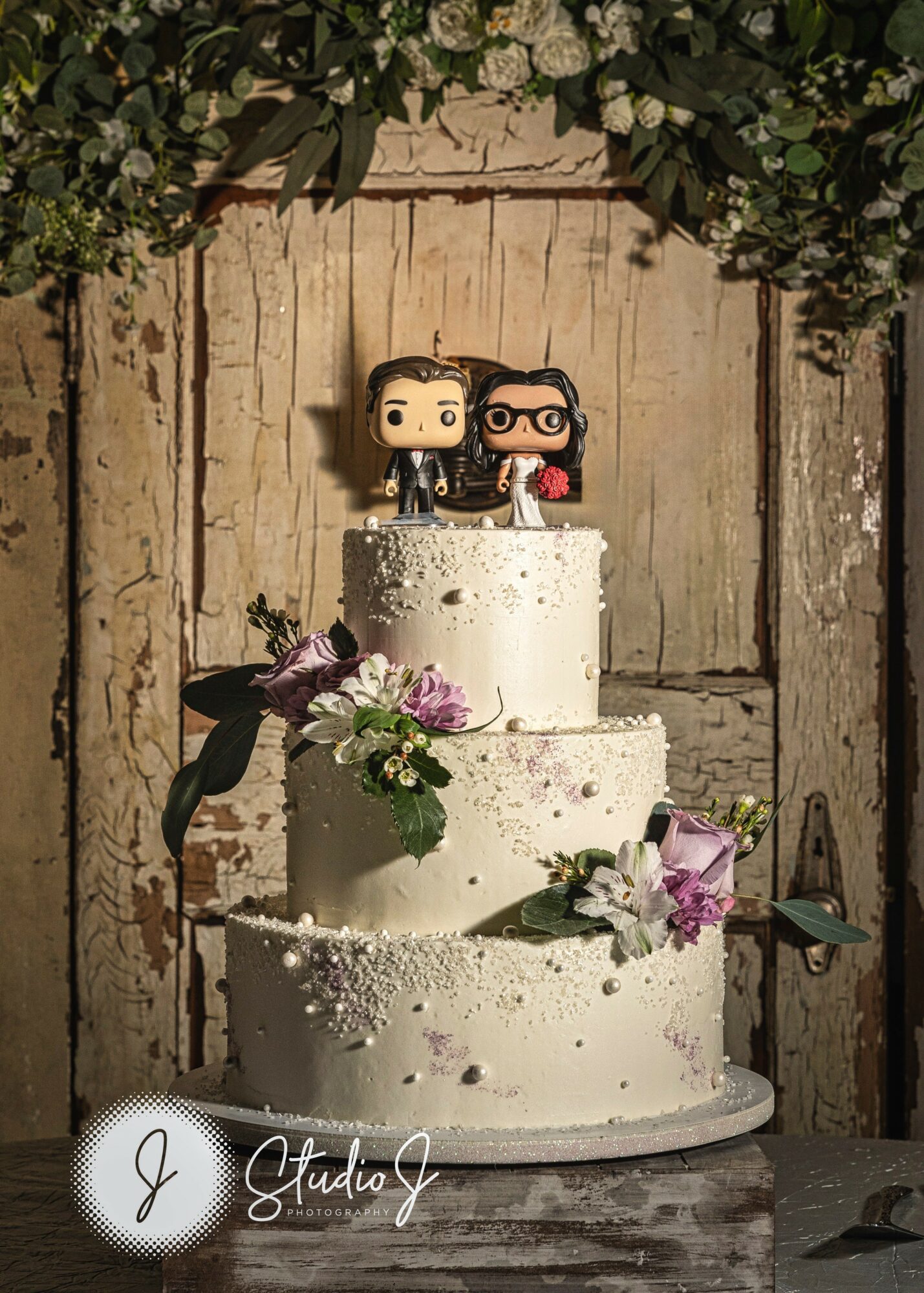 Three-tiered wedding cake with floral decorations and figurines on top, against a rustic wooden background.