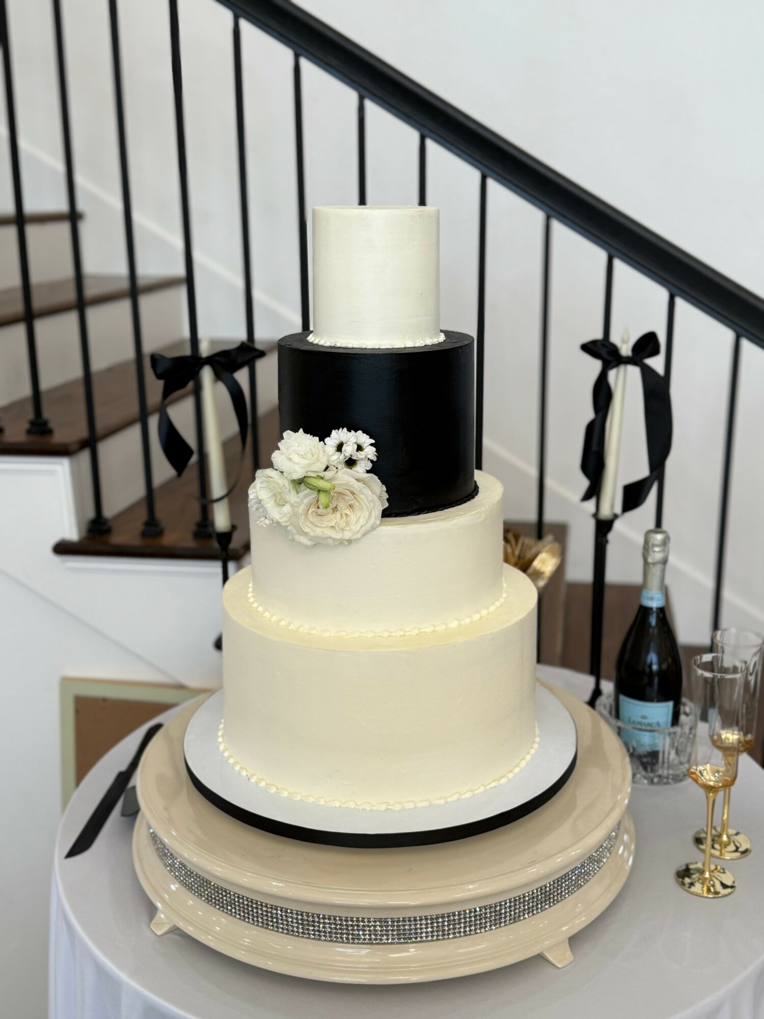 Four-tiered cake with black and white design, decorated with flowers, on a table with champagne bottles and glasses.