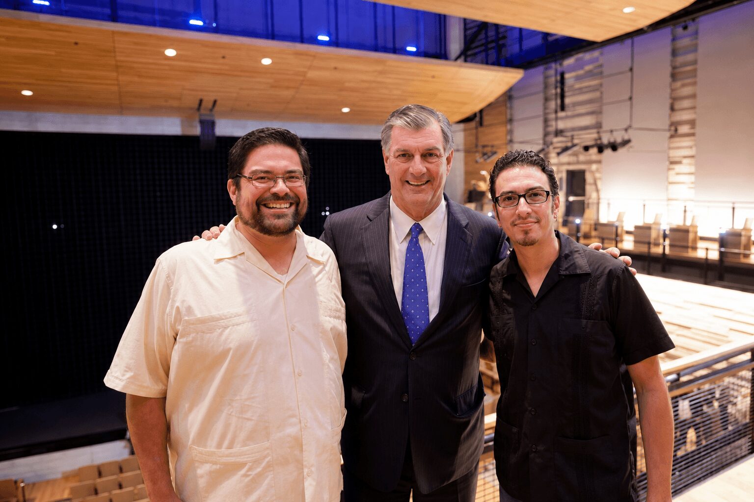 Three men standing together indoors, smiling, with a stage and seating area in the background.