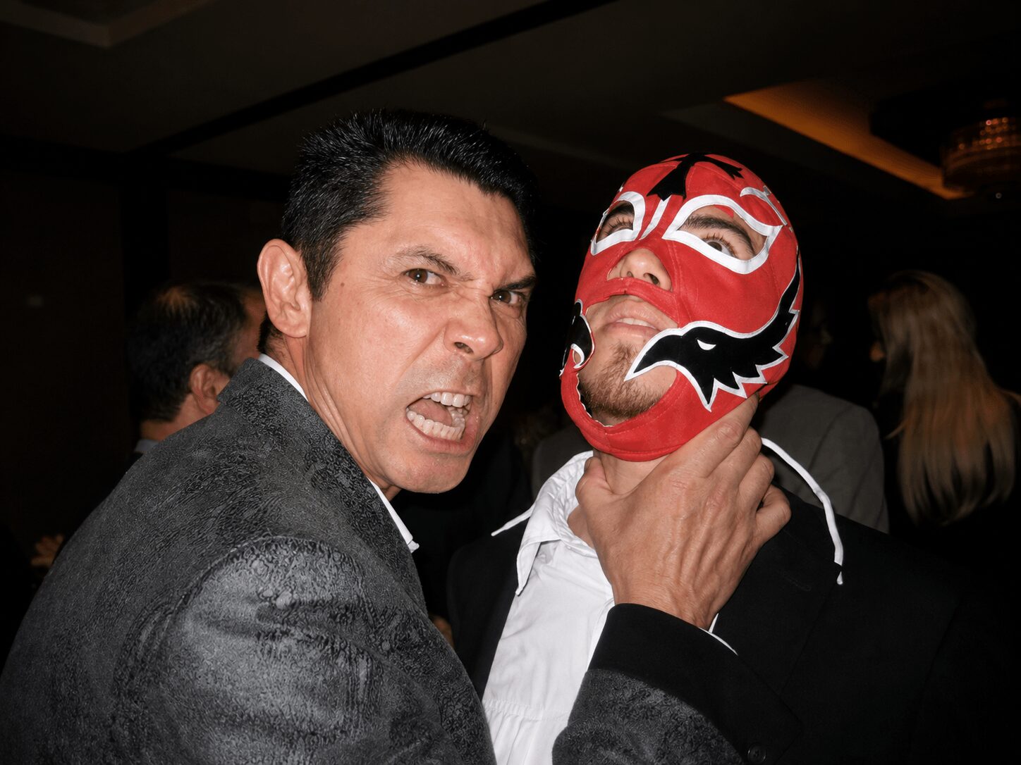 Two men at a party, one with a serious expression and the other wearing a red and white mask, in a dimly lit room.