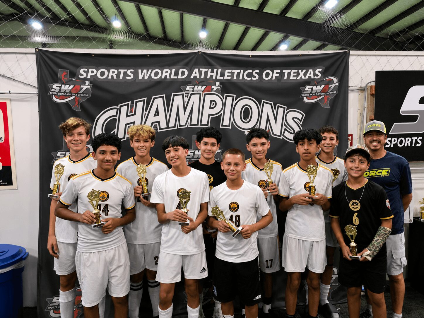 Group of young athletes in white uniforms holding trophies, standing in front of a banner that reads 'Champions' at Sports World Athletics of Texas.