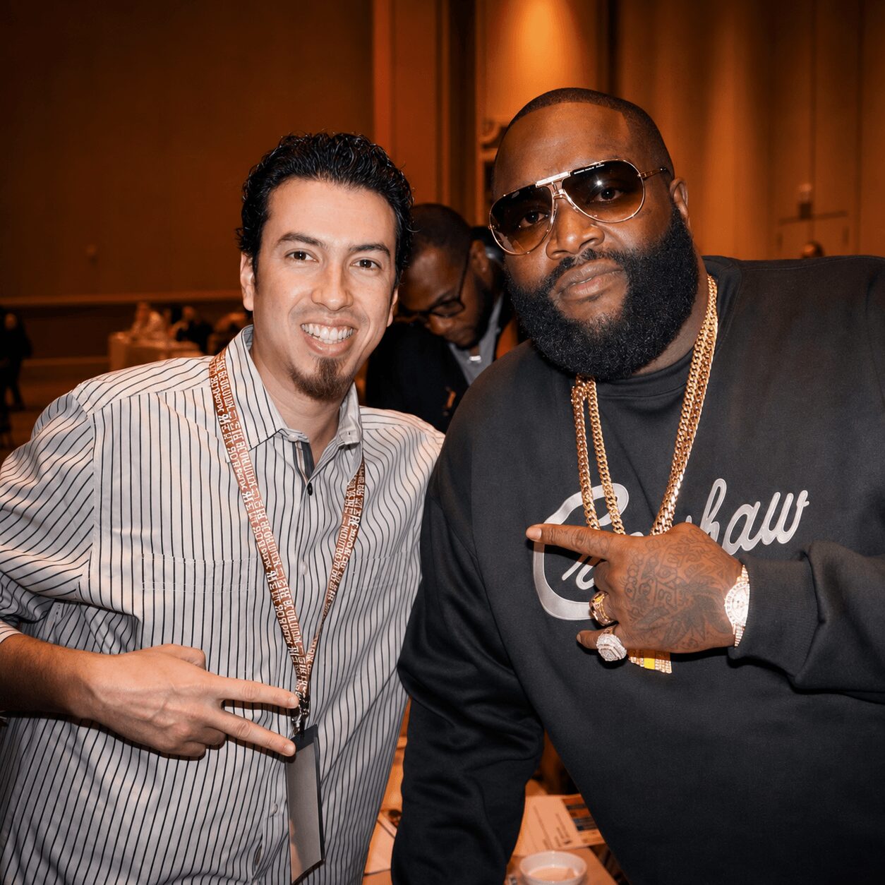 Two men posing together indoors, one wearing sunglasses and gold chains, the other smiling with a striped shirt, both making hand gestures.