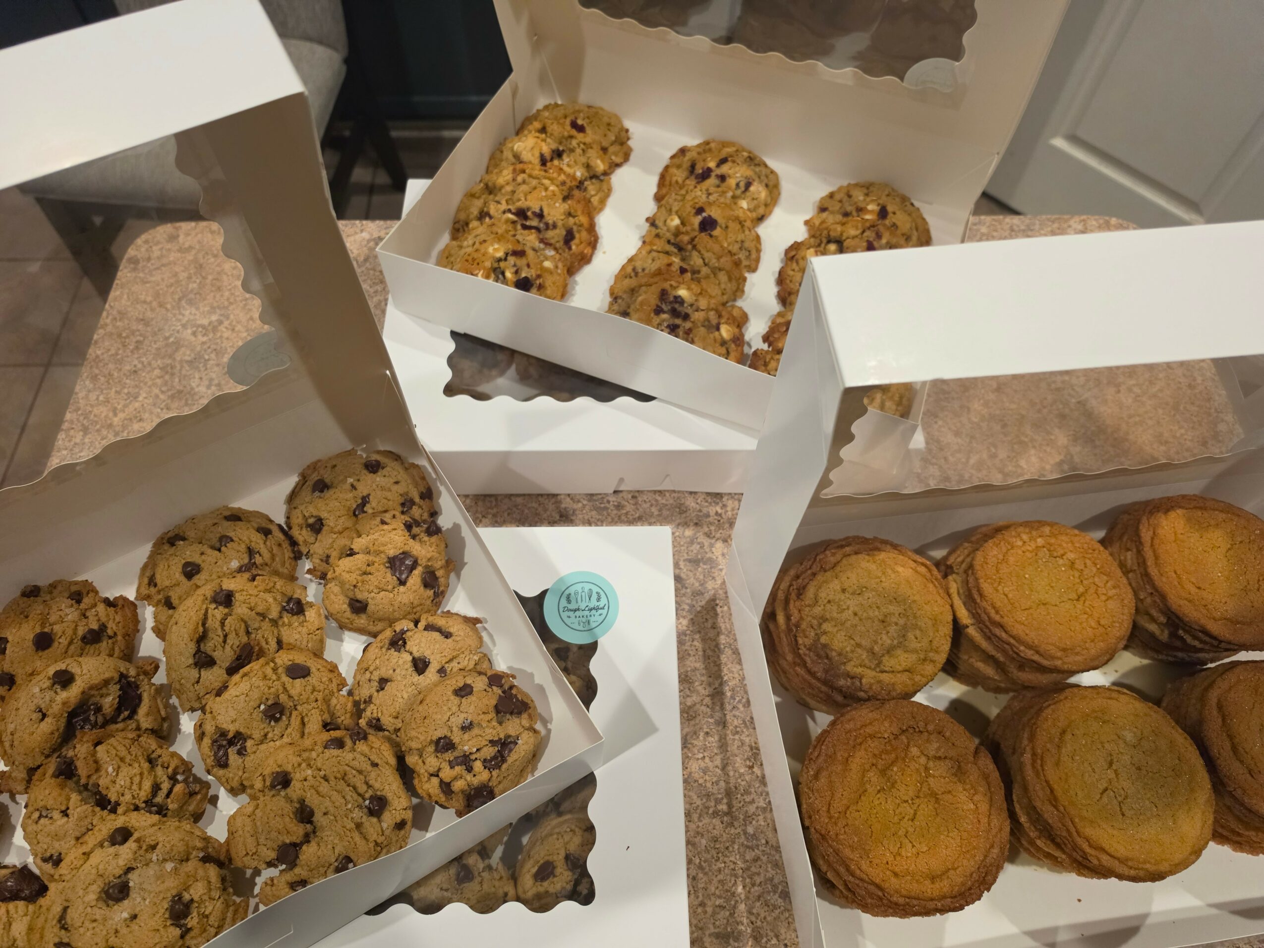 Three open boxes of baked goods on a table, containing cookies and muffins, with a blue sticker on one box.