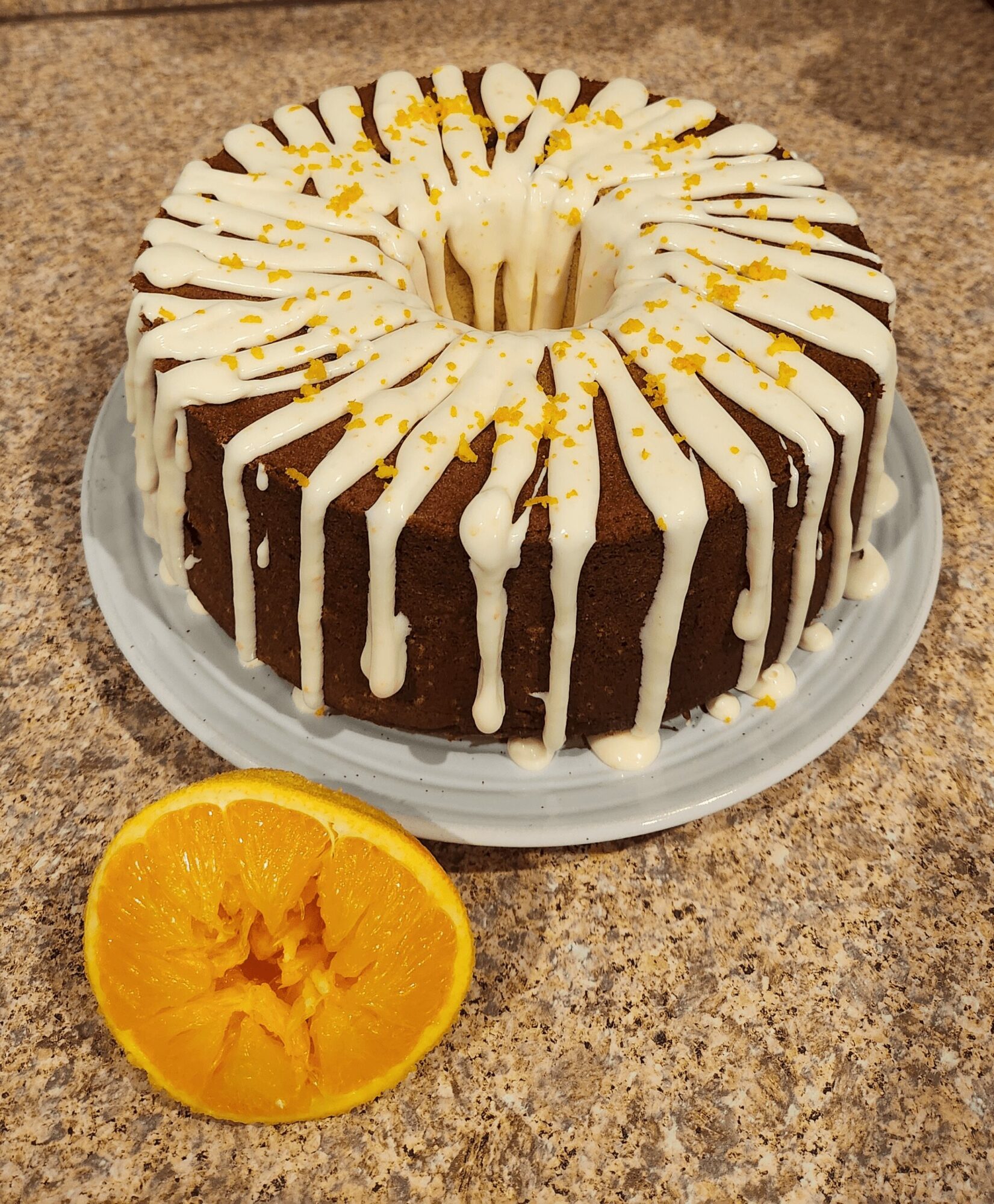 Chocolate cake with white icing drizzle and yellow sprinkles on a white plate, with half an orange in front, on a brown countertop.