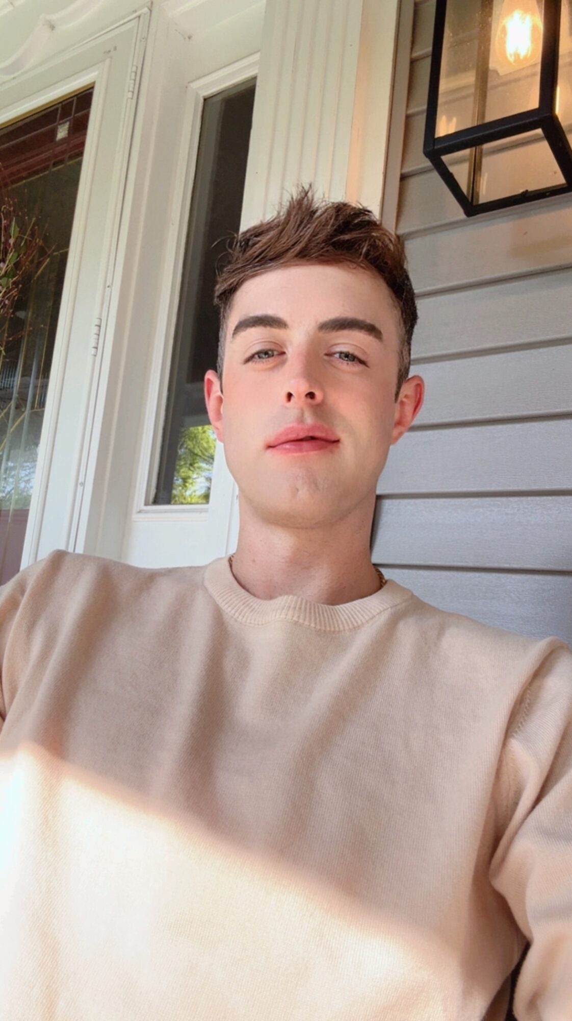 Young man with short hair wearing a beige sweater, sitting outside near a house with a window and a lantern, sunlight on face.