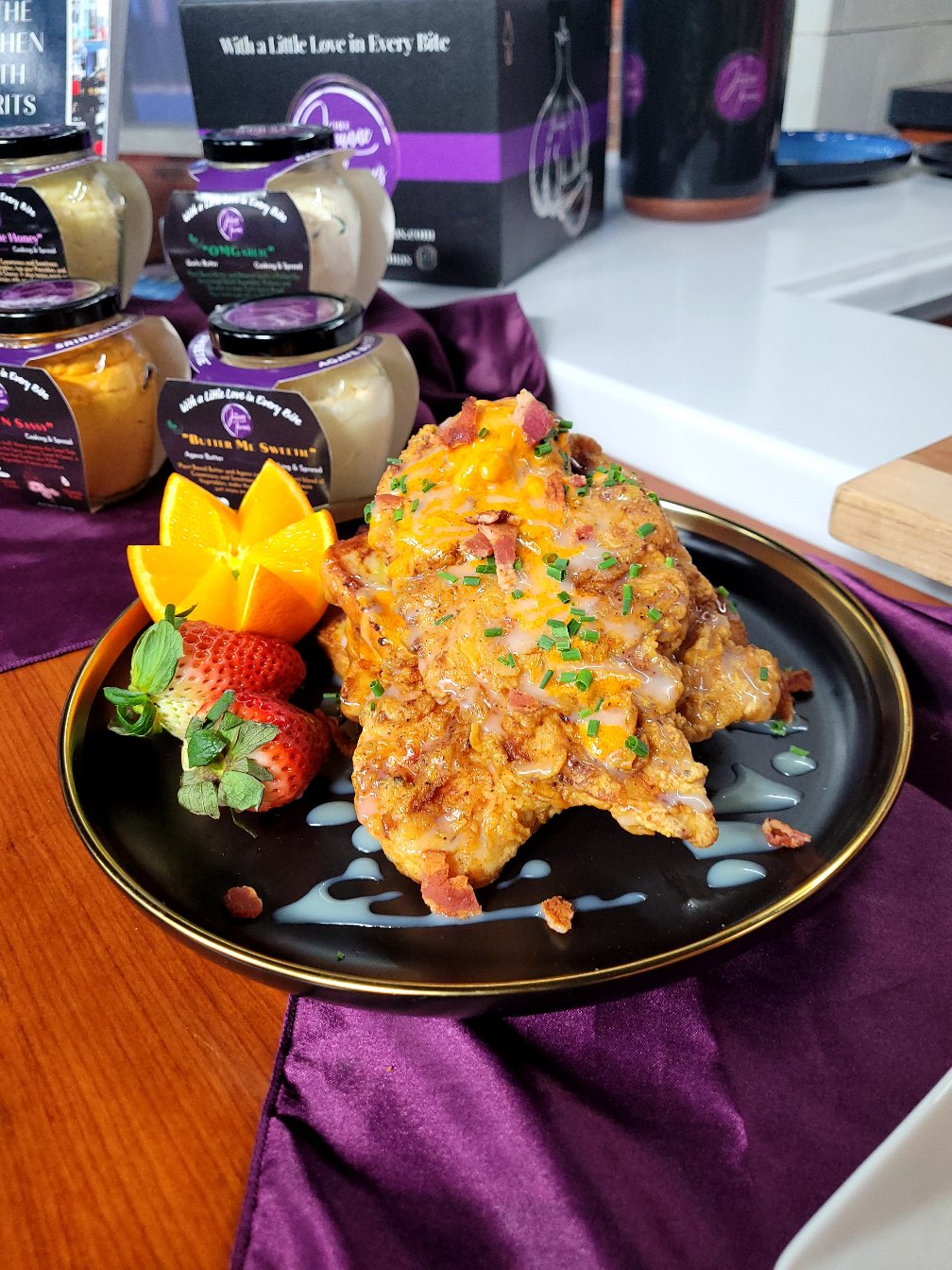 Fried chicken piece garnished with chopped herbs, served with strawberries, yellow flower, and sauce on a black plate.