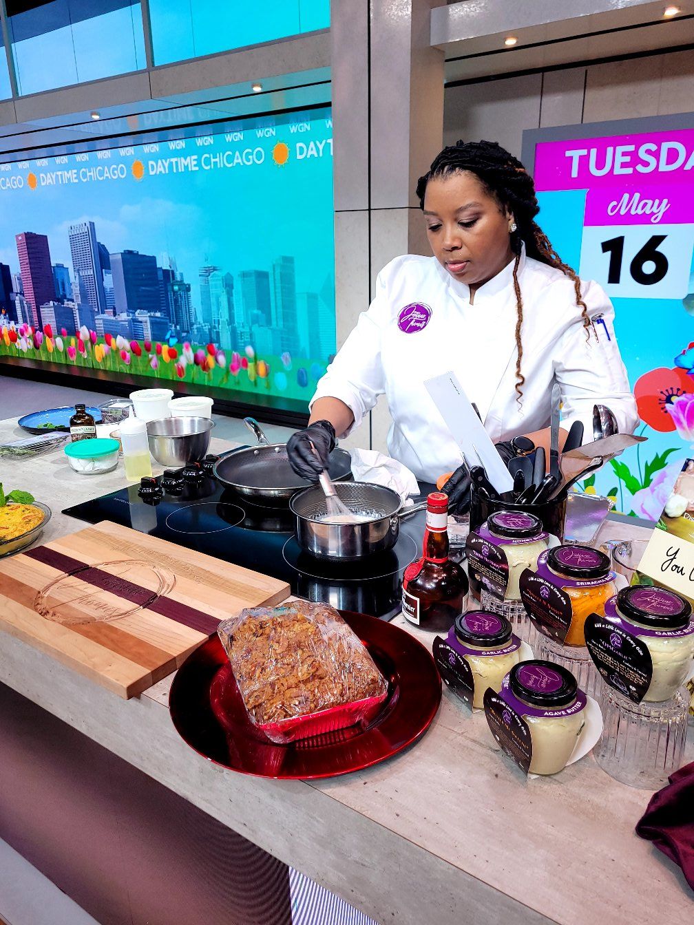 Woman in white apron preparing food at a table with desserts and drinks, in a modern indoor setting.