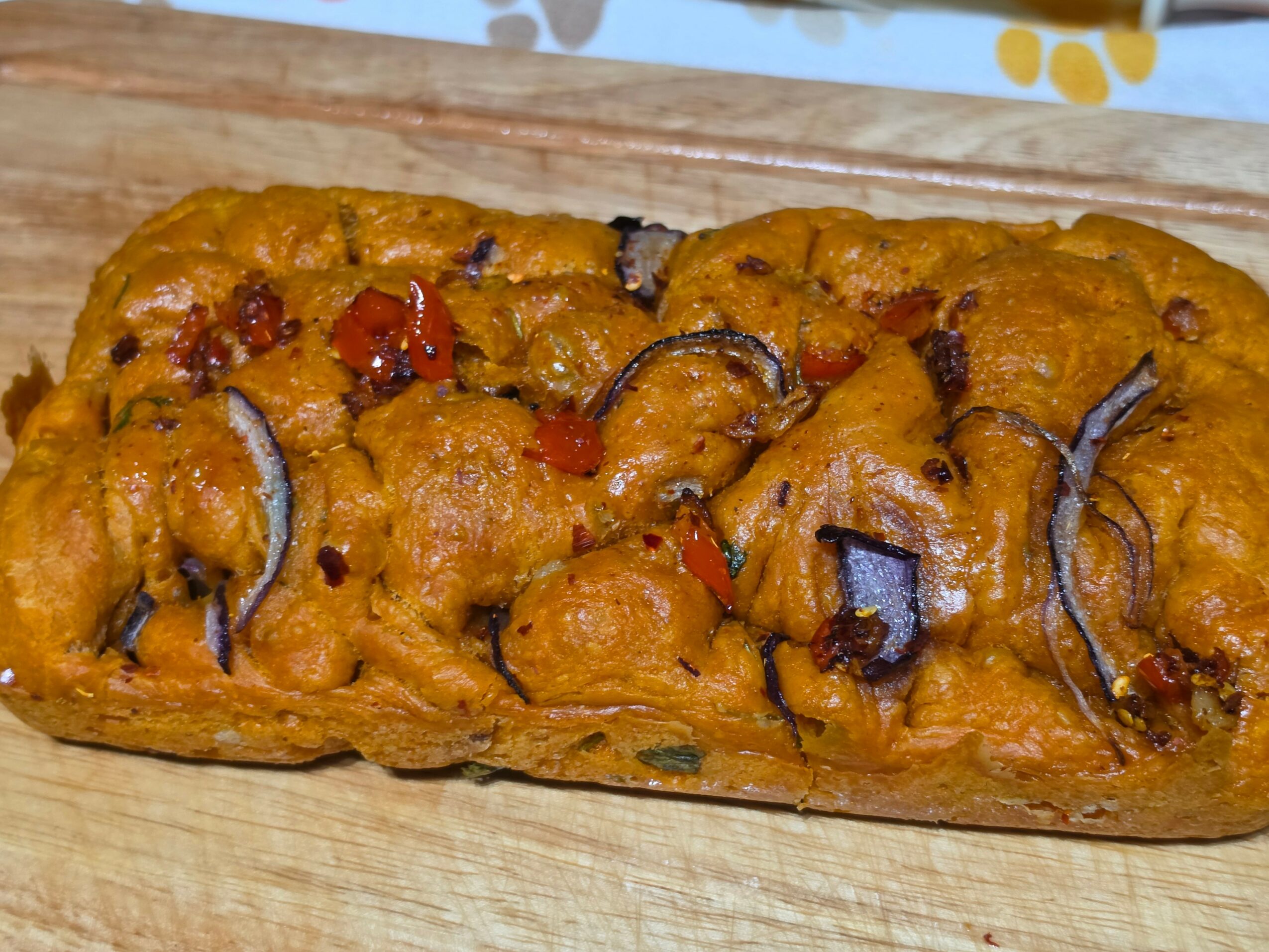 Baked bread topped with vegetables and herbs, garnished with sliced red peppers and herbs, on a wooden surface.
