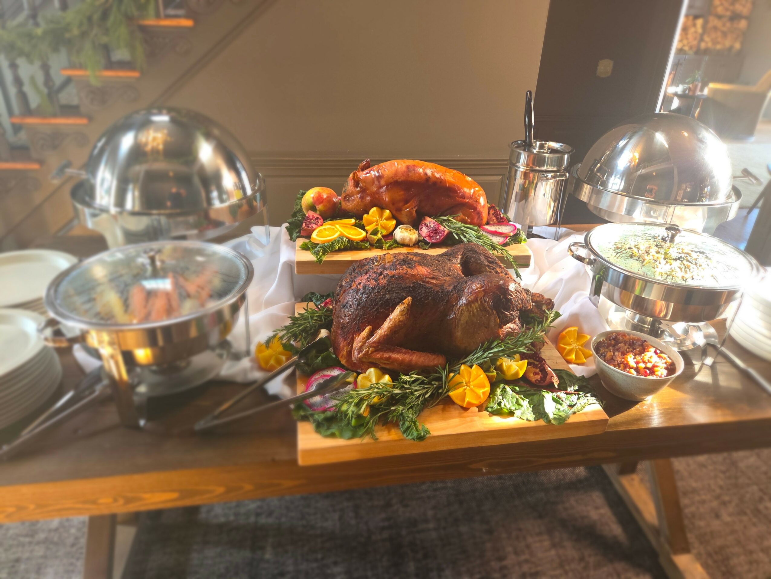 Roast turkey with stuffing, vegetables, and garnishes on a wooden table, with serving dishes and utensils around.