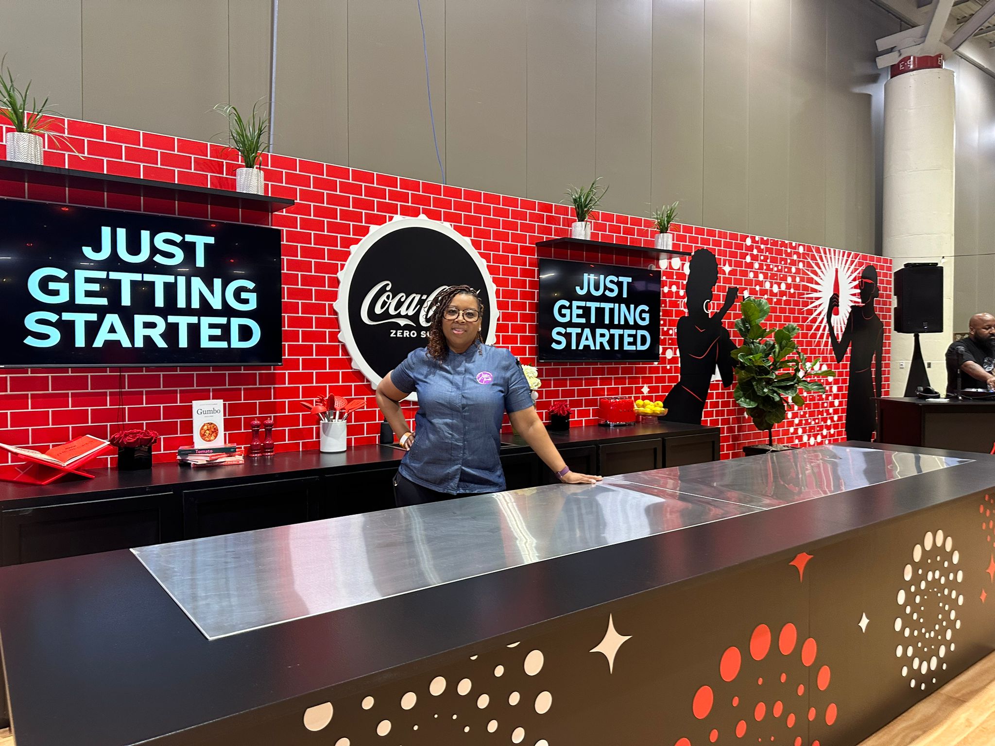 Woman standing behind a counter in front of a red brick wall with Coca-Cola logos and signs that say 'Just Getting Started'.