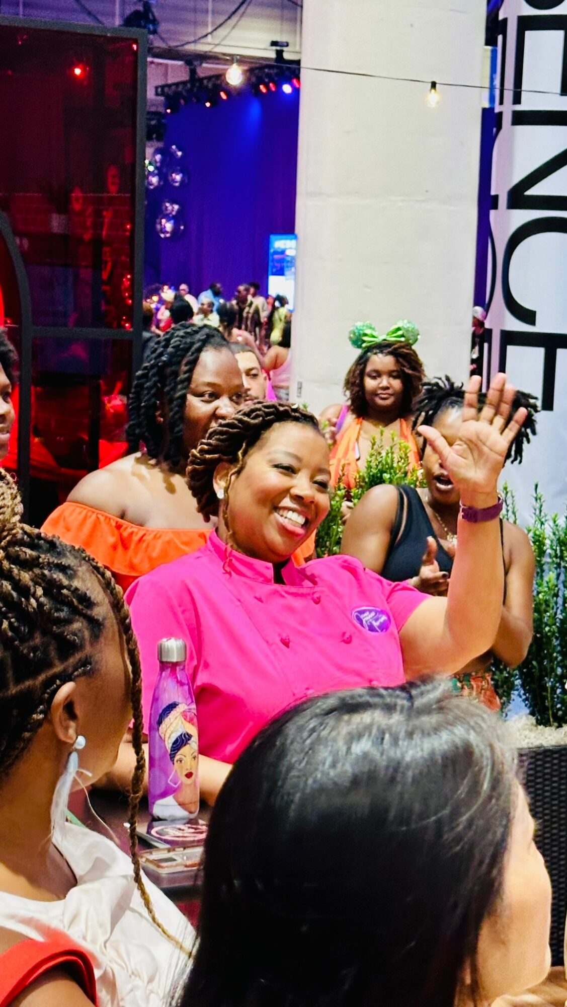 Group of smiling women and girls at an indoor event, some waving and wearing colorful clothing and accessories.