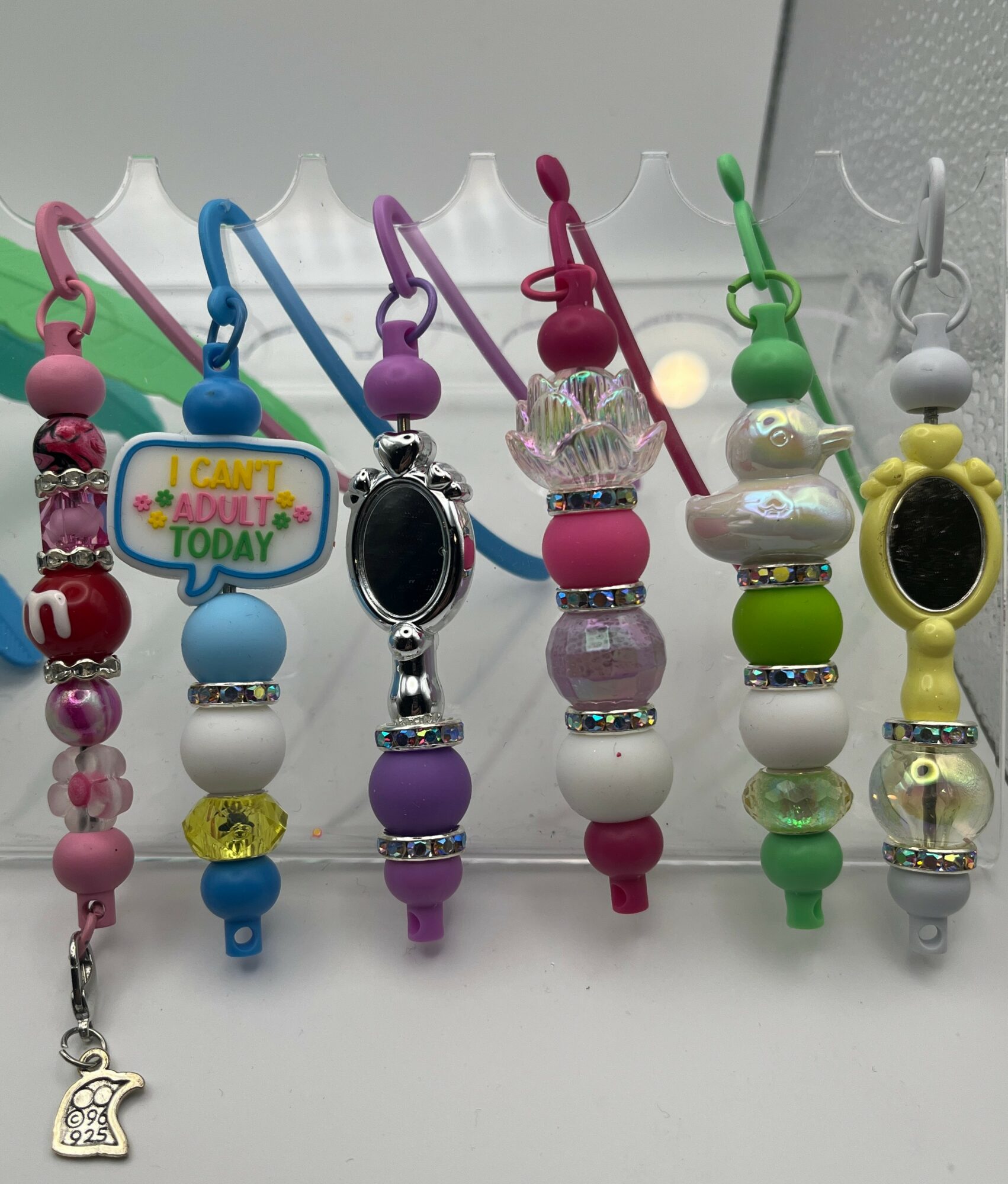 Six colorful pacifier clips with beads and watches, arranged in a row on a white surface.