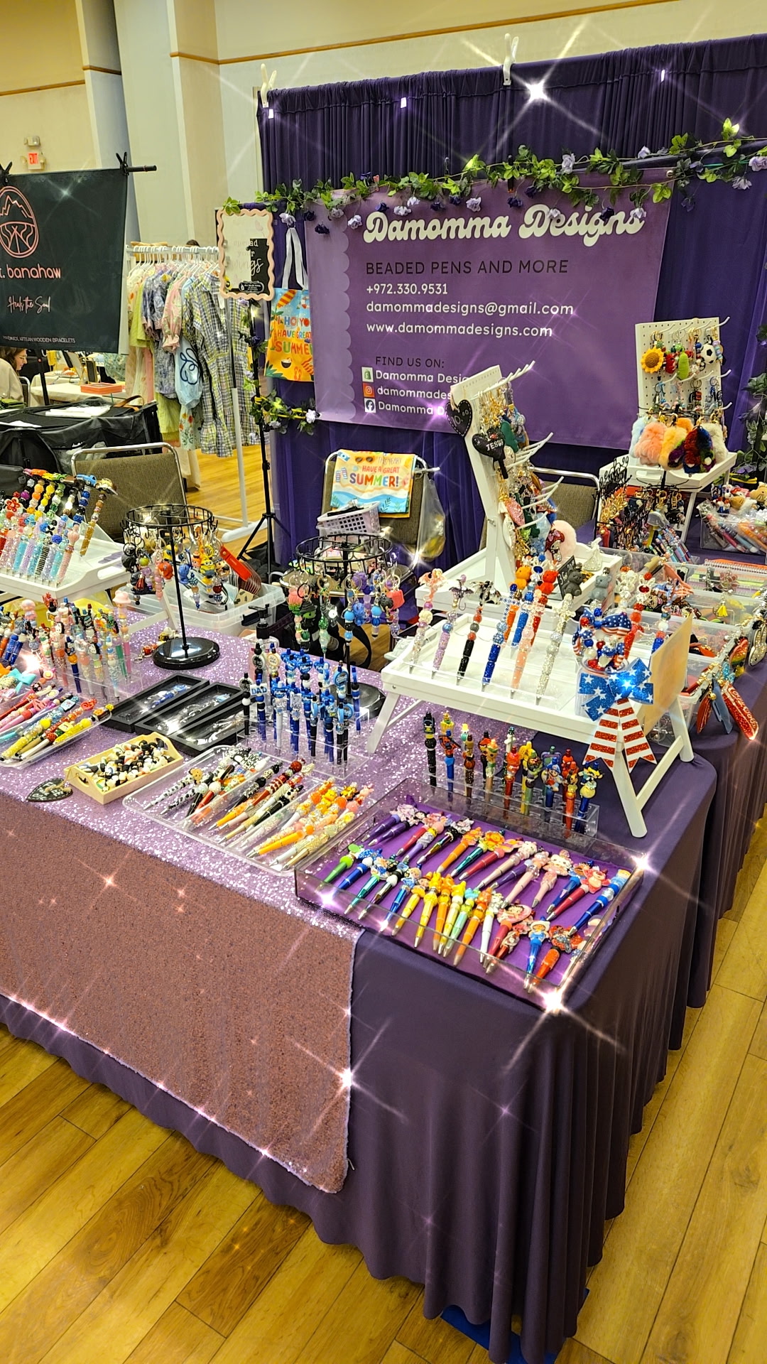 Table with colorful jewelry and accessories at a craft fair, purple backdrop with text, and various displayed items.